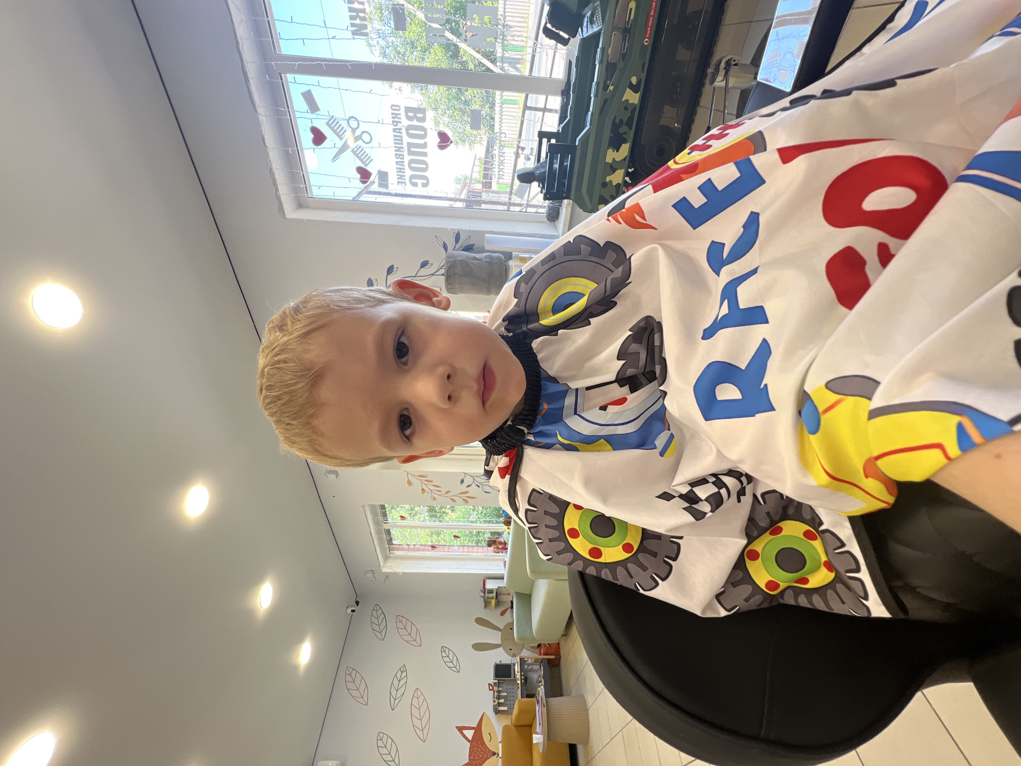 A young Caucasian boy with blond hair sits in a black barber chair, wearing a white cape with a racing car theme. The cape features images of car tires, checkered flags, and the word "RACE" in large blue letters. He is looking directly at the camera with a neutral expression.
The background suggests a brightly lit children's hair salon. The walls are white and decorated with cartoonish illustrations of leaves and a fox. To the left, a yellow sofa and a low coffee table are visible. Further back, a light green sofa and a toy kitchen set are present.
A large window with decorative cutouts of scissors and hearts runs along the back wall, offering a view of trees and a street. Some lettering on the window appears to be in Russian. A green toy military vehicle sits on a shelf or display in front of the window. The ceiling has recessed lighting fixtures. The overall atmosphere appears to be that of a child-friendly establishment. The presence of a children's hair salon in Bataysk, Russia is consistent with the visual cues.