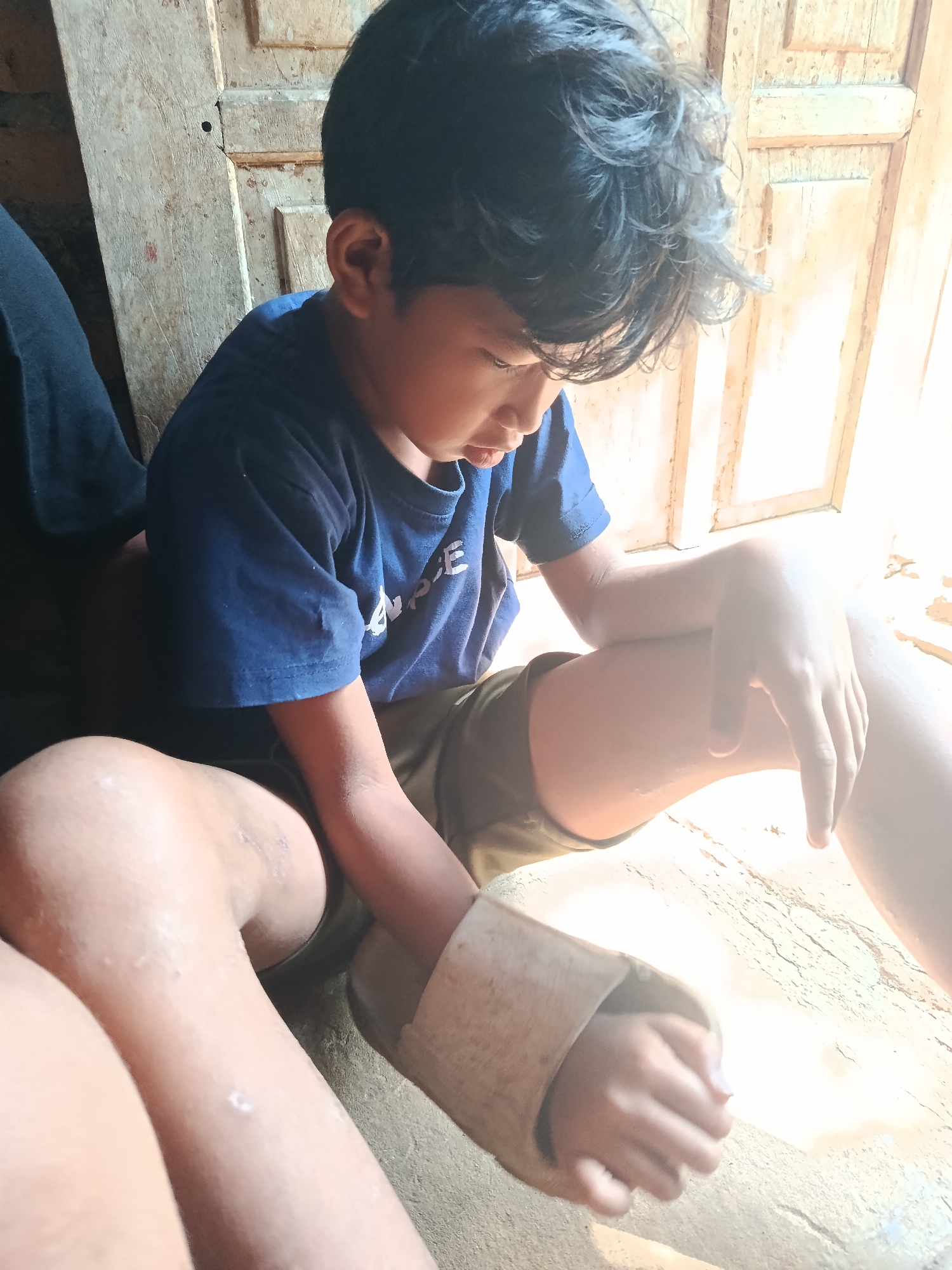 This media file depicts a young boy sitting on a wooden floor, likely indoors. The boy is wearing a dark blue t-shirt and olive-green shorts. He is looking down, with his head slightly bowed, and his left arm is resting on his knee. His right arm is bent, with his hand resting on his thigh. A sandal is visible on his left foot. The background shows a wooden door or wall, with light streaming in from the right, suggesting it is daytime and possibly sunny. The overall scene has a casual and relaxed atmosphere. There is some faint white text visible on the boy's t-shirt, but it is not clearly legible. The location context is Karangmojo, Indonesia.