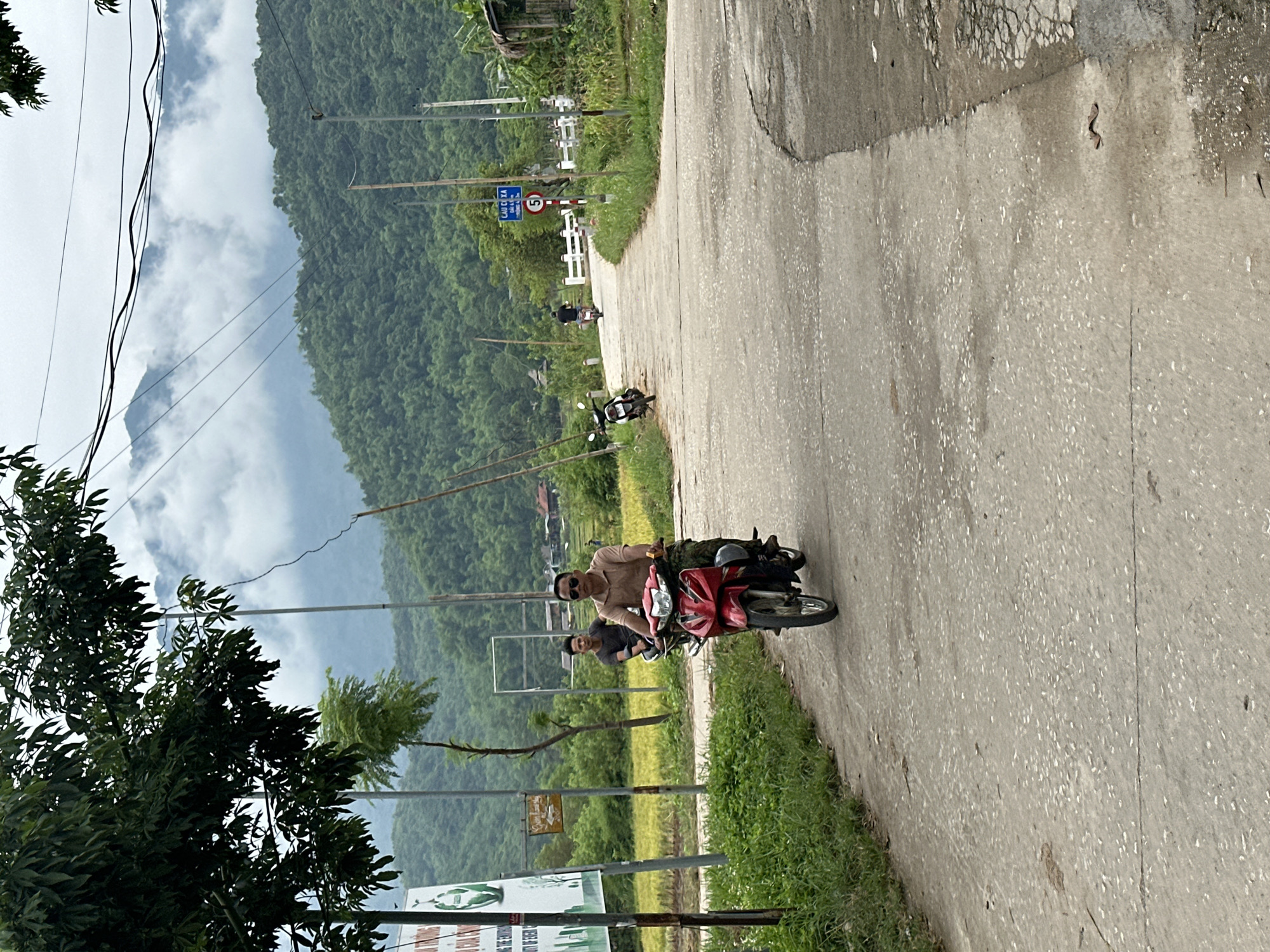 The image depicts a rural road in Mai Chau, Vietnam, with lush green hills and a cloudy sky in the background. A man is riding a red motorcycle on the paved road, wearing a light brown shirt and camouflage pants. Another man is riding a motorcycle behind him. To the left of the road, there is a field of yellow crops, a signpost with Vietnamese text and an image of a recycling symbol, and a tree. Further down the road, there is a speed limit sign indicating 5 and a white bridge. The road is lined with grass and vegetation. The scene appears to be captured during the daytime.