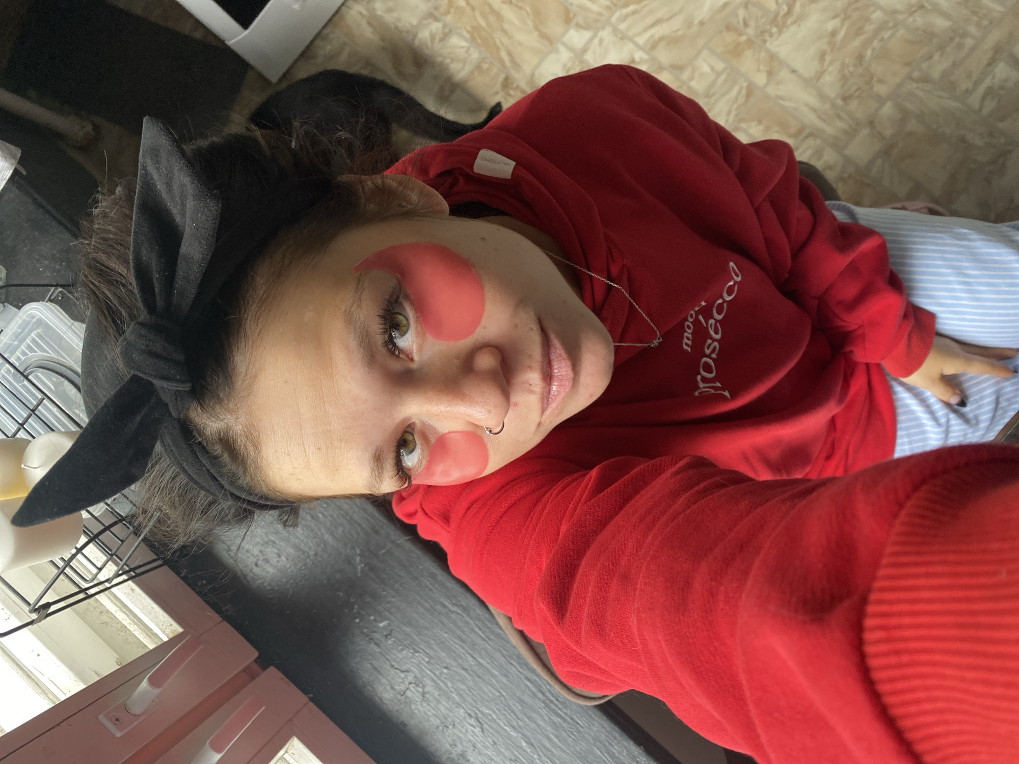 A woman with light brown hair is taking a selfie indoors. She is wearing a red hooded sweatshirt with the word "prosecco" embroidered in white on the front. She has pink under-eye patches on, a nose ring, and a black headband with a bow on top of her head. The woman is seated, and her legs are visible from the knees down. She is wearing light blue and white striped pants. The background of the image includes a window with white trim and pink handles, a black surface, and a tiled floor. The overall setting appears to be a domestic interior, potentially a kitchen or living area. There is no indication of the image being taken in Chaikovsky, Russia.