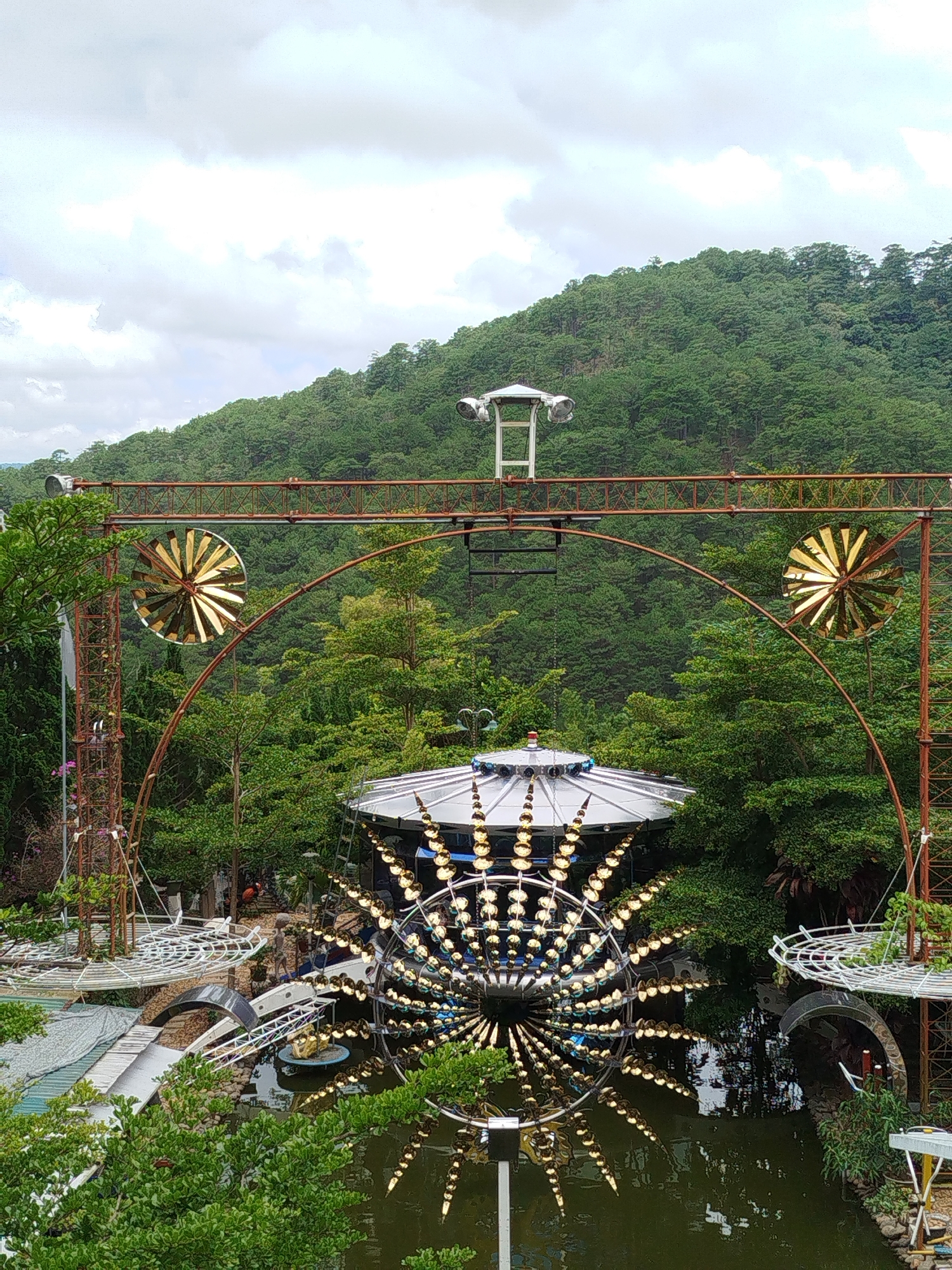 The image shows an outdoor art installation in Da Lat, Vietnam, likely at the Da Lat Flower Park or a similar botanical garden known for its artistic displays. In the foreground, a large, circular kinetic sculpture with reflective gold-colored vanes is partially submerged in a dark body of water. Above this, a domed structure with mirrored surfaces and illuminated elements is visible, possibly part of a pavilion or attraction. Extending over the scene is a rusty metal archway supporting two golden, pinwheel-like decorative elements. In the background, a densely forested hillside rises under a cloudy sky, characteristic of the mountainous terrain surrounding Da Lat. The overall scene depicts a whimsical and artistic arrangement set within a lush, natural environment.