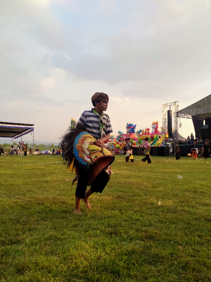 The image captures an outdoor cultural performance on a vibrant green grassy field in Glonggong, Indonesia, under an overcast sky suggesting late afternoon. The central subject is a young male dancer, positioned slightly right of center, mid-air with his left foot raised. He wears a horizontally striped white and blue t-shirt, dark pants, and a green patterned scarf around his neck. Dark markings are visible on his face. Attached to his waist is an elaborate, two-dimensional, colorful horse effigy with a dark, flowing mane, indicating a traditional Indonesian dance performance. He is barefoot.
In the background, a large black stage structure with audio speakers is visible on the right. To the left of the stage, several large, brightly colored inflatable play structures, including a prominent robot figure and various cartoon animals, are present. Further left, a white and blue canopy tent is erected. Scattered individuals, including other performers in similar attire and spectators, are dispersed across the field and near the background structures. White debris is visible on the grass in the foreground.