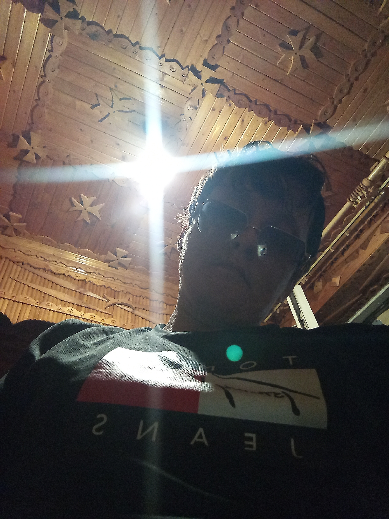 A low-angle, indoor, first-person perspective selfie shows a person wearing sunglasses and a dark t-shirt with a logo. The t-shirt logo features a white rectangle with a red stripe and the word "JEANS" printed below. The background consists of a ornately carved wooden ceiling and walls, suggesting an interior with traditional or rustic decor. A bright light source, possibly a camera flash or overhead lighting, creates a lens flare that streaks across the image, obscuring some details of the background and the person's face. The setting appears to be a residential or commercial space with a distinct architectural style. There is no information available to determine the specific location in Vsevolozhsk, Russia.