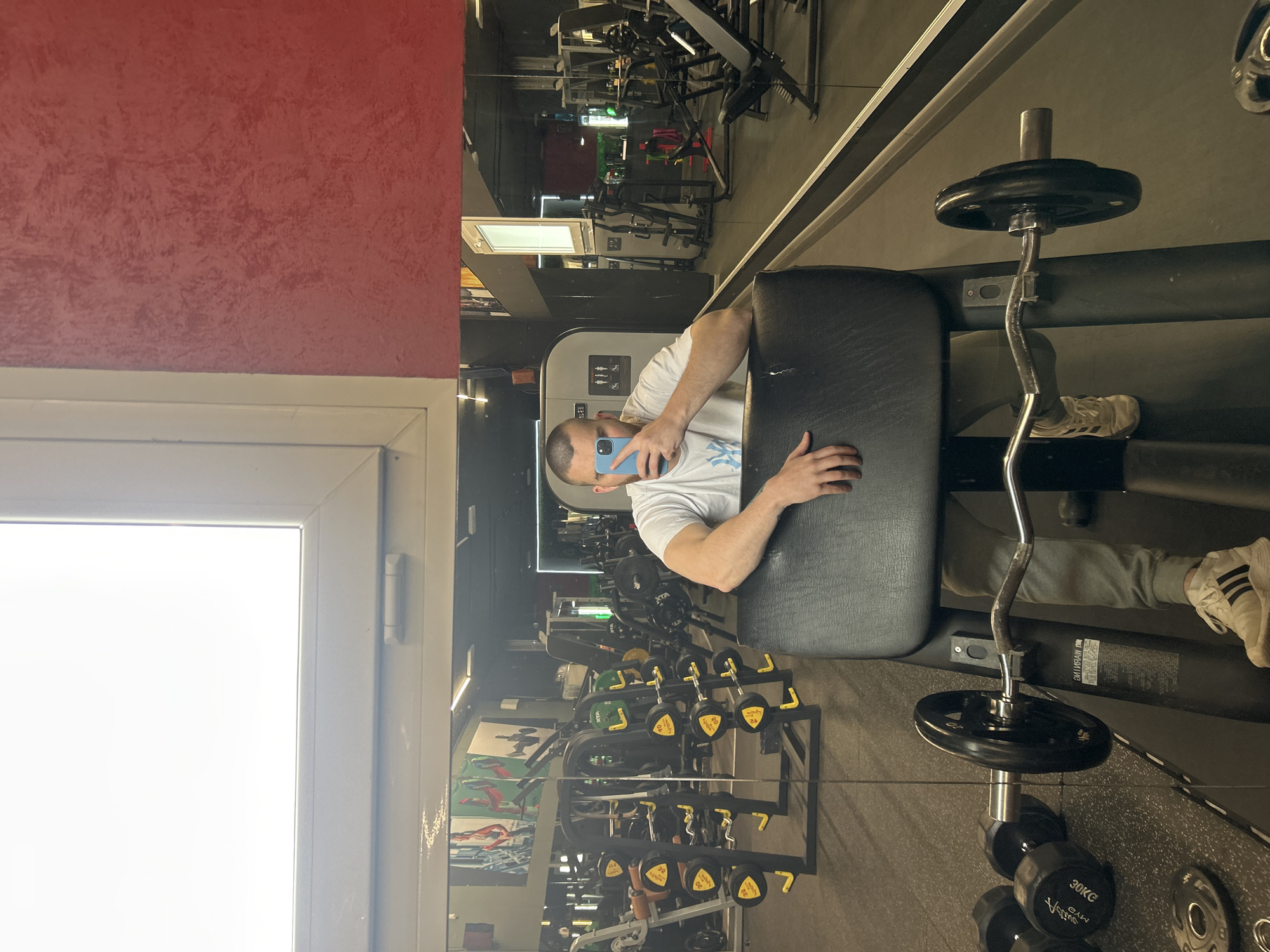 A man in a white t-shirt, holding a blue phone, is reflected in a mirror in what appears to be a gym. The reflection shows workout equipment, including weights and machines. The man is sitting on a workout bench next to a barbell with weights loaded on it. A window and red wall are visible on the left side of the image. The location is not specified.