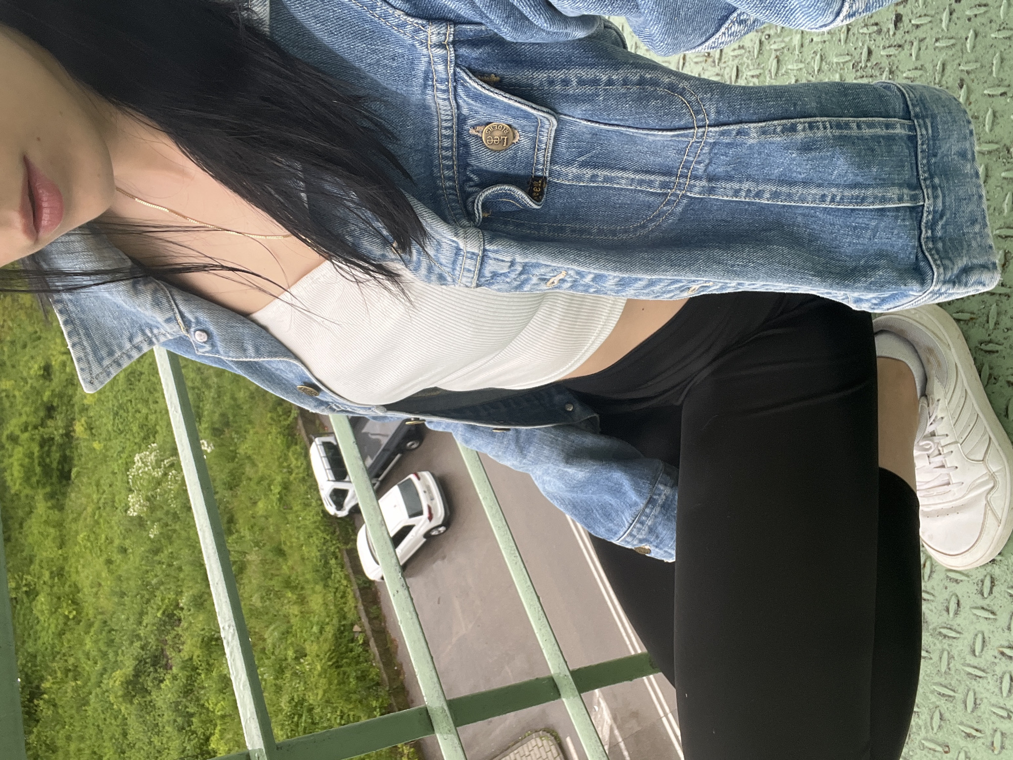 An overhead, slightly tilted shot captures a person from the chest down, positioned on an elevated green metal platform with a railing, looking down at the ground below. The person wears a light blue denim jacket, a white ribbed crop top, black leggings, and white sneakers with white socks. A thin gold-colored chain necklace is visible around their neck, and the lower part of their face shows lips with light pink lipstick. Long, dark hair cascades over their shoulders.
Notable details on the denim jacket include a metal button on a chest pocket flap, which clearly reads "LEE RIDERS," and a small "Lee" fabric tag attached nearby. The green metal platform has a textured, diamond-plate pattern.
Below the railing, an asphalt road with white markings is visible, along with two white cars parked on the side. Beyond the road, there's a lush green area covered in grass and bushes, with scattered white wildflowers. The scene appears to be set during daytime, with soft lighting suggesting either an overcast sky or early morning/late afternoon.