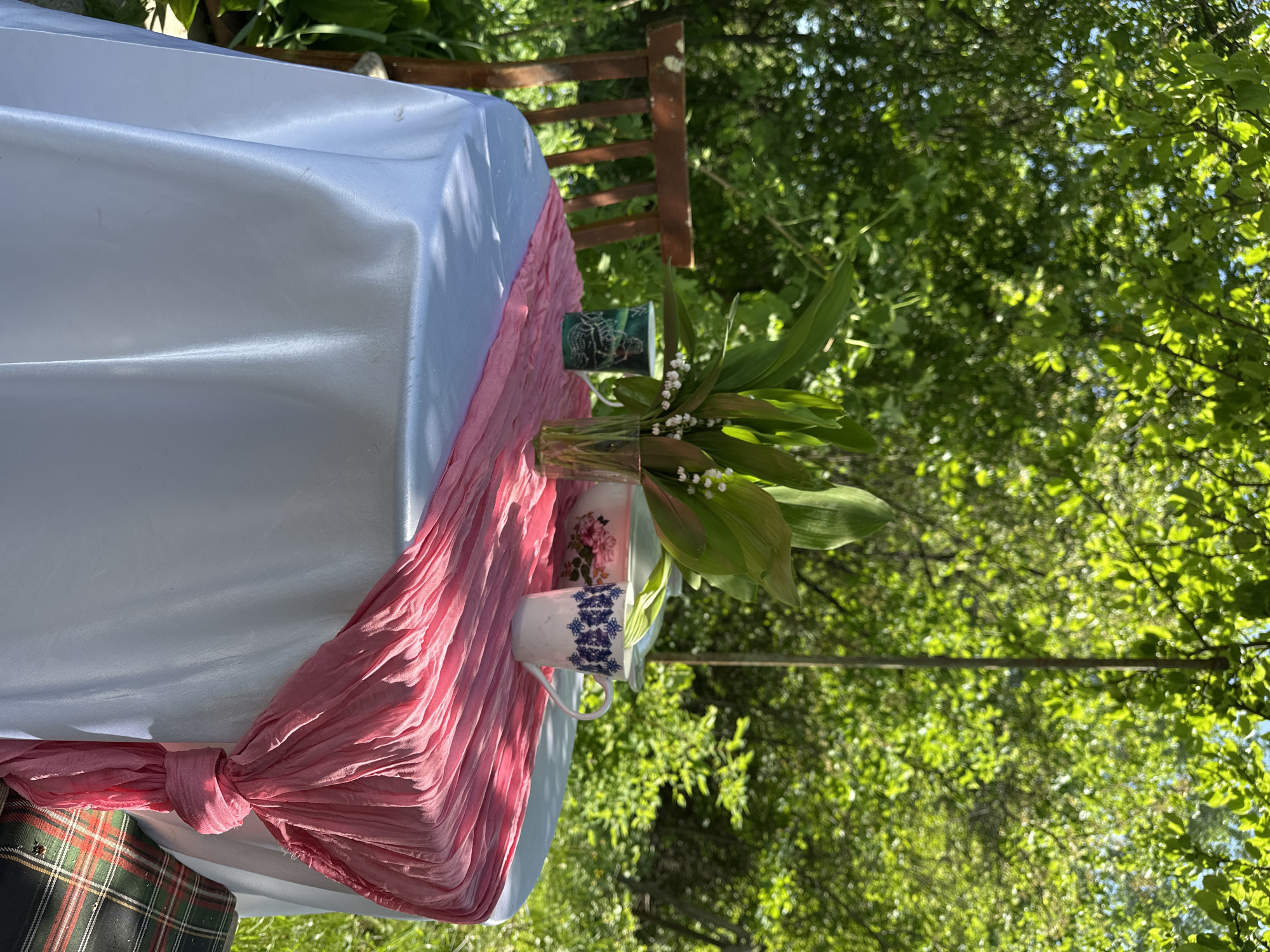 The image presents an outdoor setting, likely a garden. A table is draped with a white cloth and adorned with a pink runner. On the table, there are a few cups, and a vase with lily of the valley. The background is filled with green foliage and trees, suggesting a natural environment. Based on the prompt, the location is Chaikovsky, Russia.