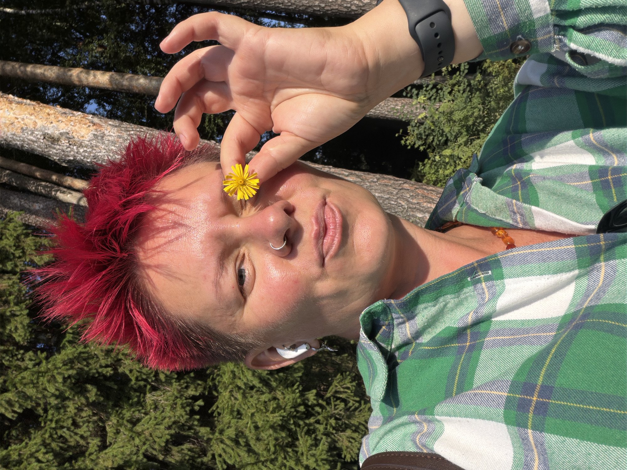 A woman with spiky, bright red hair holds a small yellow flower in front of her right eye. She is wearing a green and white plaid shirt, a necklace made of amber beads, and a smartwatch. She also has a nose ring and an earbud in her left ear. The background shows a forest with tall trees and green foliage, suggesting an outdoor setting in Chaikovsky, Russia. The lighting indicates it is daytime, likely a sunny day.