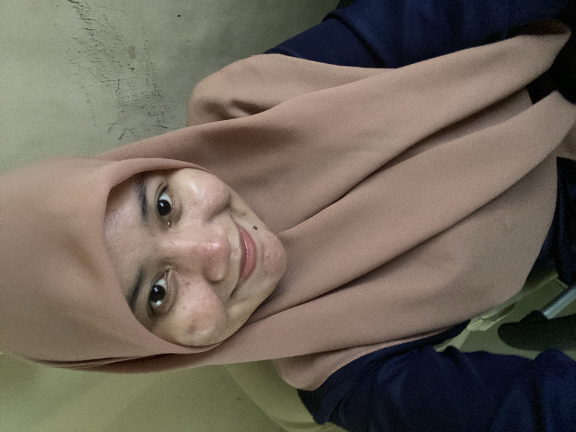 A close-up, selfie-style, medium shot captures a young woman wearing a light brown hijab and a navy blue shirt. She is positioned in the center of the frame, looking directly at the camera with a subtle smile. Her face, illuminated by direct light, shows some skin blemishes. The background is a textured, pale yellow wall with some dark smudges. The image appears to be taken indoors, possibly in a residential setting within the city of Banjarbaru, Indonesia.