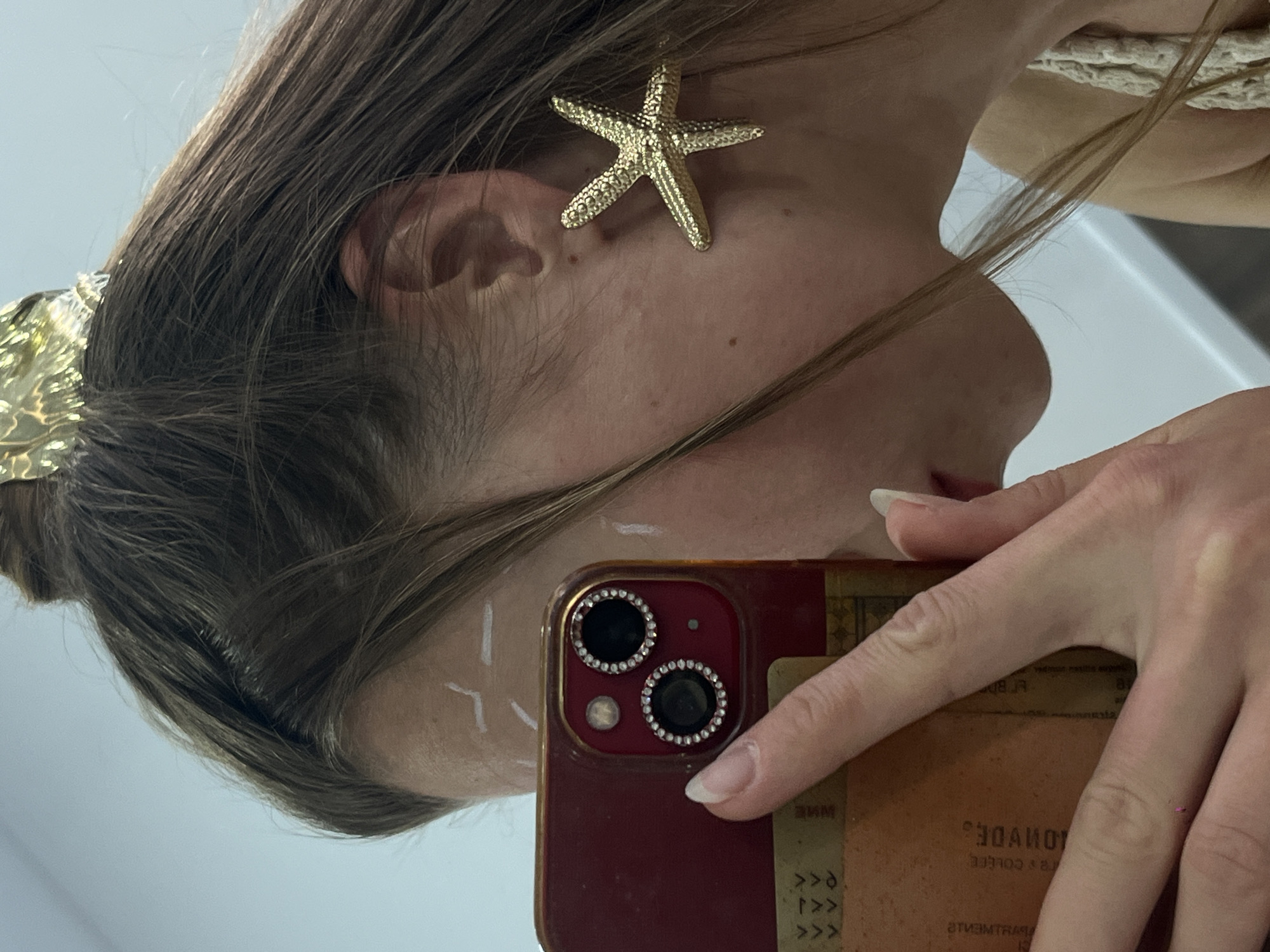 A close-up, angled, selfie-style photograph shows a person with brown hair wearing a gold starfish earring. The person's face is partially visible, and there is white paint or makeup applied in a geometric pattern near their temple. The person is holding a smartphone with a decorated camera lens area, and a weathered-looking card or attachment on the back. The background is indistinct but appears to be an indoor space with a reflective surface, possibly a mirror. It is impossible to determine any details about the city of Budva or any connections to Montenegro based on the image provided.