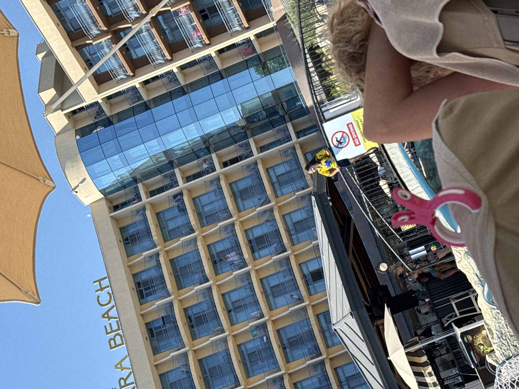 The image depicts a sunny outdoor scene, likely at a resort, with a large building in the background. The building, a multi-story hotel, has a beige facade and blue glass windows, with balconies on each floor. Above the hotel, a large tan umbrella is visible, partially obscuring the blue sky. In the mid-ground, a railing separates an elevated walkway from a pool area. A sign with a "No Diving" symbol is prominently displayed on the railing. A young person in a yellow shirt stands near the sign. In the foreground, a person's legs and arm are visible, suggesting they are reclining in a lounge chair. A pink object, possibly a toy or accessory, is in the lower right quadrant of the image. In the lower left, a bar or food stand is partially visible, with umbrellas and seating. The overall impression is a vacation setting. Based on the building signage that reads "ARA BEACH," this location is likely the Ara Beach hotel in Nesebar, Bulgaria.