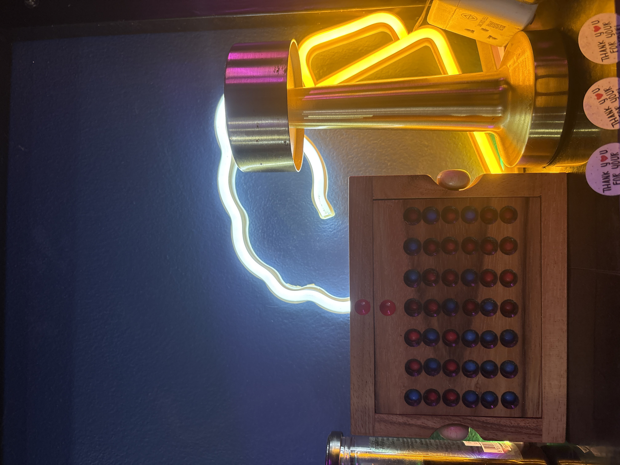 The image depicts a close-up view of a dimly lit interior space. The primary elements are two neon signs, one shaped like a hand in white light and the other with a yellow-orange geometric design. These are mounted on a dark blue wall. Adjacent to the neon signs is a wooden game board with multiple round colored pieces (red and blue) embedded within. The scene also includes elements such as stickers that read "THANK YOU FOR YOUR" and fragments of bottles. The context is suggestive of an establishment or venue, although the specific location within Baan Bang Rak, Thailand, cannot be determined from the image alone.