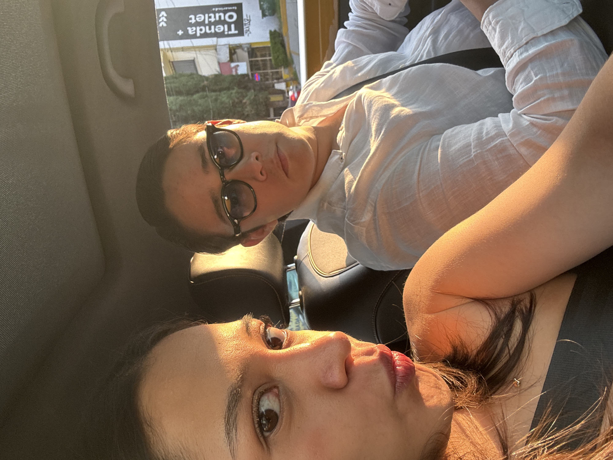 Two people are in a car; one is wearing a white, long-sleeved shirt, and glasses with black frames; the other is wearing a black top. The background indicates they are in Puebla, Mexico, near a "Tienda + Outlet." The car has a gray interior.