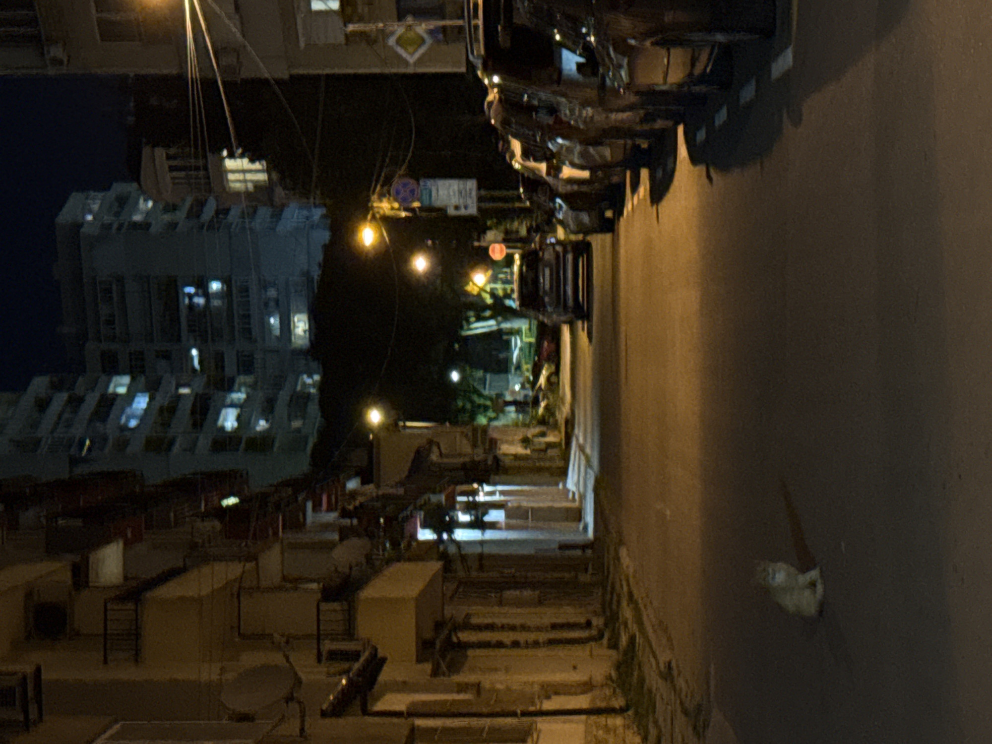 A nighttime street scene in an urban area is presented. The road is dark and adjacent to a row of parked vehicles. A multi-story building stands in the background. Illumination from street lamps casts a glow on the surroundings. A low-rise structure sits on the left. A blurry, white shape rests on the street.