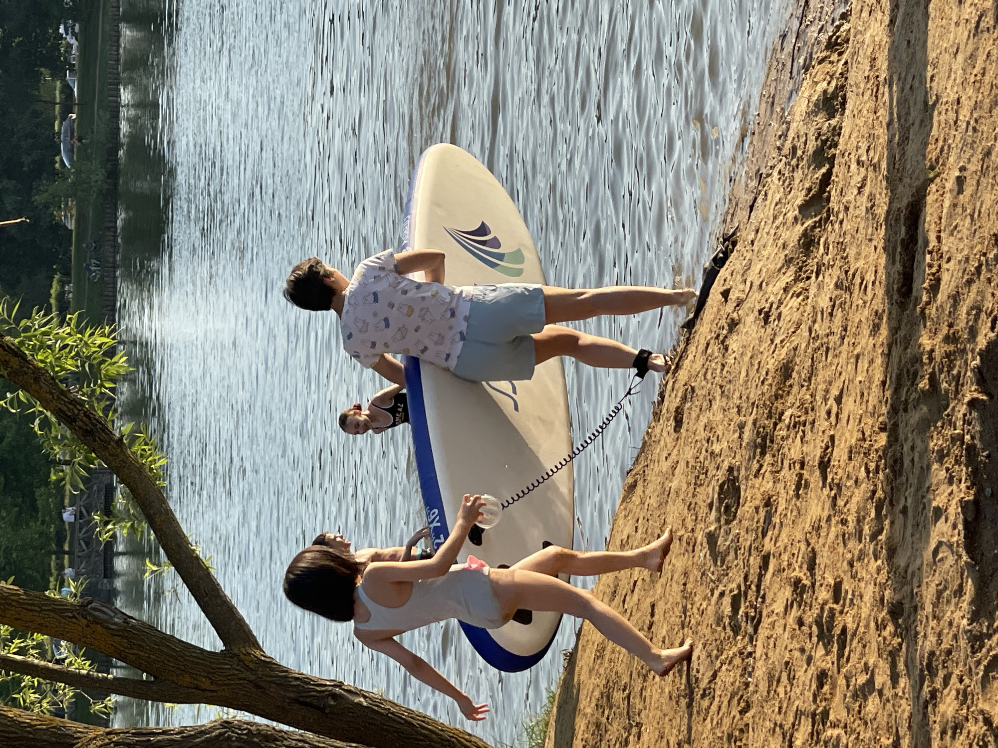 The scene depicts a recreational waterfront area in Moscow, Russia, featuring a sandy bank leading to a large body of water. Four individuals are present in the foreground and midground, engaged in or preparing for water activities.
In the left foreground, a young female with short, dark hair, wearing a light grey one-piece swimsuit, walks barefoot on the textured, sunlit sandy slope towards the water. Adjacent to her, a second individual, viewed from behind, with short dark hair, is dressed in a white short-sleeved t-shirt patterned with small cartoon characters and light blue shorts. This person stands with their right foot partially submerged in the water, carrying a white inflatable stand-up paddleboard (SUP). The paddleboard displays blue and green graphic designs and the text "2'X6'". A black coiled leash connects the individual's right ankle to the paddleboard.
Further in the water, behind the first female, a third person, shirtless, is partially visible. To the far right in the water, a fourth young female with reddish-brown hair tied up, wearing a dark swimsuit, smiles while looking left towards the group.
A large tree trunk with green foliage partially frames the left side of the scene. The body of water, likely a river or lake, exhibits gentle ripples. In the distant background across the water, a treeline, a light-colored path, and parked vehicles are visible, characteristic of a public recreational zone along a large waterway within the city.