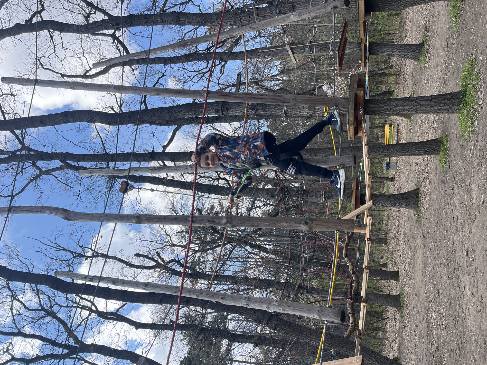 A young boy is participating in a treetop adventure park. He is suspended in the air between trees on a rope obstacle course. He is wearing a patterned jacket, dark pants, and athletic shoes. He is holding onto a red rope. The course appears to be constructed with wooden platforms and ropes strung between trees. The trees are mostly bare of leaves, suggesting it is not summer. The sky is visible between the branches and is partly cloudy. The ground below is covered in dirt and dead leaves. The scene depicts an outdoor recreational activity in a park-like setting, likely in the city of Sofia, Bulgaria.