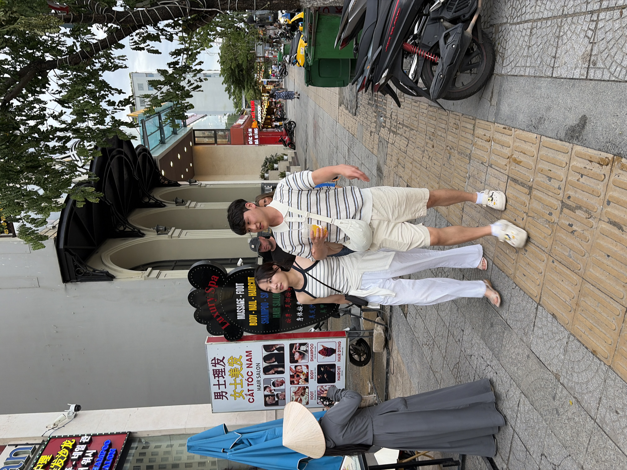 The image depicts a street scene in Da Nang, Vietnam. A young Asian couple is walking on a sidewalk. The man, wearing a striped polo shirt and beige shorts, holds a drink. The woman, in a white striped tank top and white pants, carries a small black bag. They are walking past a street vendor wearing a traditional conical hat and a grey robe, who is looking at their phone. In the background, there are shops and signs, including one for "Luxury Spa" and another advertising hair salon services with images of hairstyles. Motorbikes are parked along the street. The environment appears to be a busy urban area with a mix of buildings and pedestrian traffic.