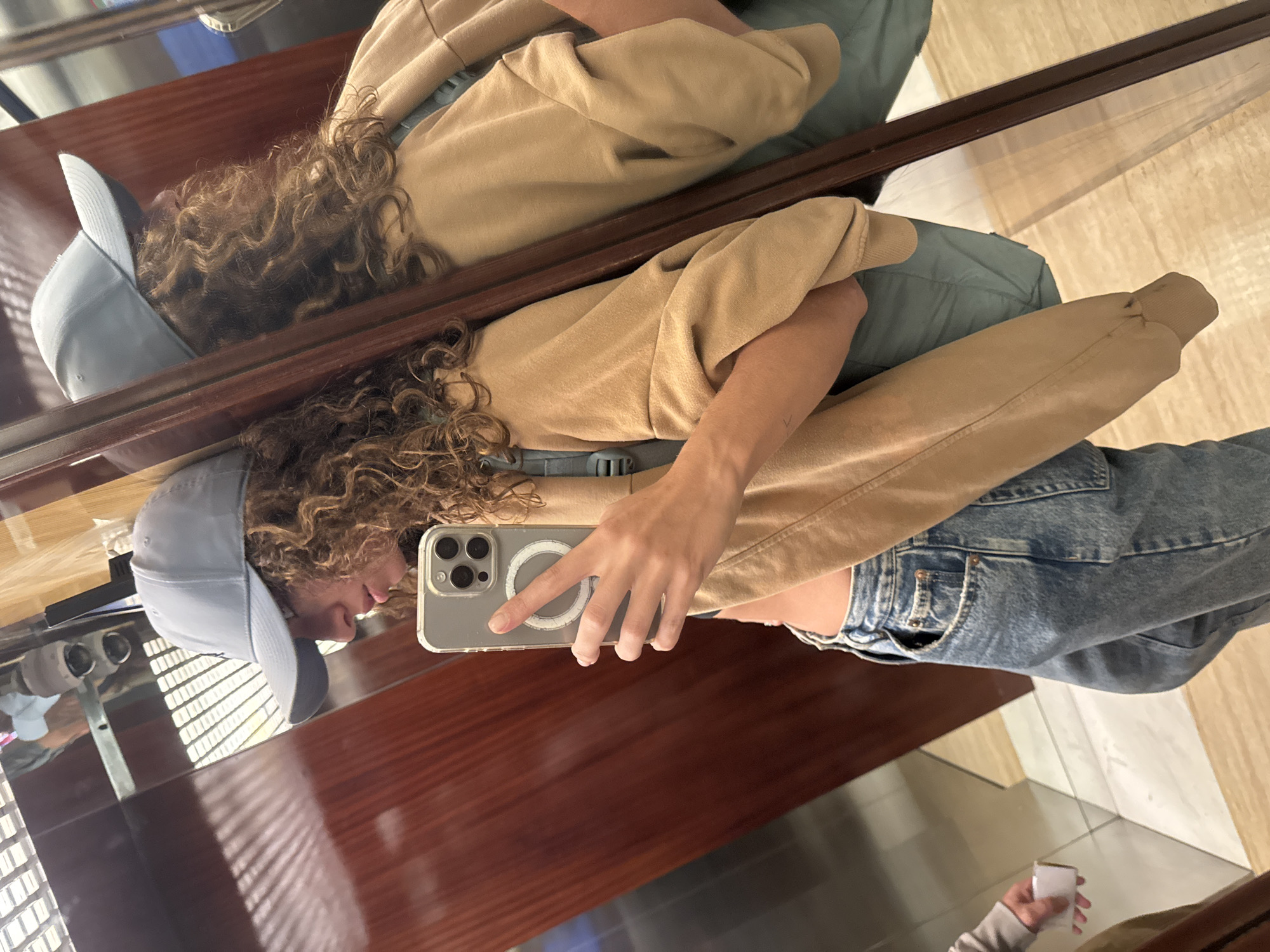 A person wearing a light blue baseball cap, a tan hoodie, and jeans is reflected in a glass surface. The person is holding a silver iPhone with a MagSafe charger. The background features a wooden surface and a light-colored tiled floor. A hand holding a small white object is visible in the reflection in the lower right corner. The location is not discernible from the provided image.
