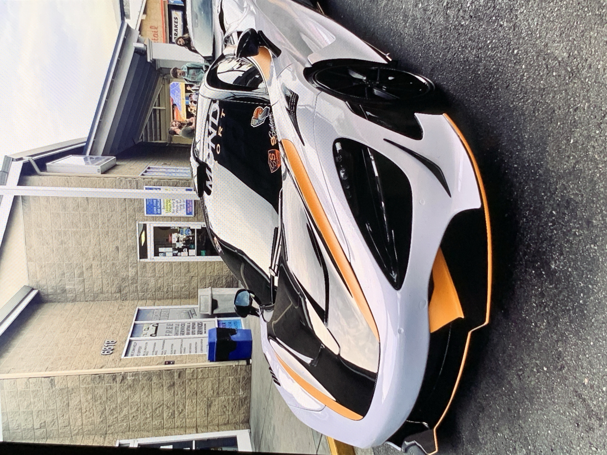 This is a medium shot, eye-level, outdoor photograph of a white McLaren 720S with black and orange accents parked on asphalt pavement. The car is positioned at an angle, facing towards the viewer's left. The front bumper is visible, with sharp orange accents and black aerodynamic elements. The headlights are integrated into the front bodywork. The car has a black roof and a black windshield banner with white text that reads "MINDSPORT". There are also two small stickers on the windshield, one of which features a stylized "SS" logo.
In the background, there is a tan brick building with the number "6818" above a window. The window displays information about services offered, including "FREE SHUTTLE SERVICE" and "AUTO REPAIR SERVICES". There are two trash cans visible near the building, one blue and one gray. To the right of the building, a covered walkway leads to an entrance with a sign that partially reads "tail" and "BRAKES". Several people are standing under the walkway, engaged in conversation. The sky is overcast, suggesting a daytime scene with diffused lighting.