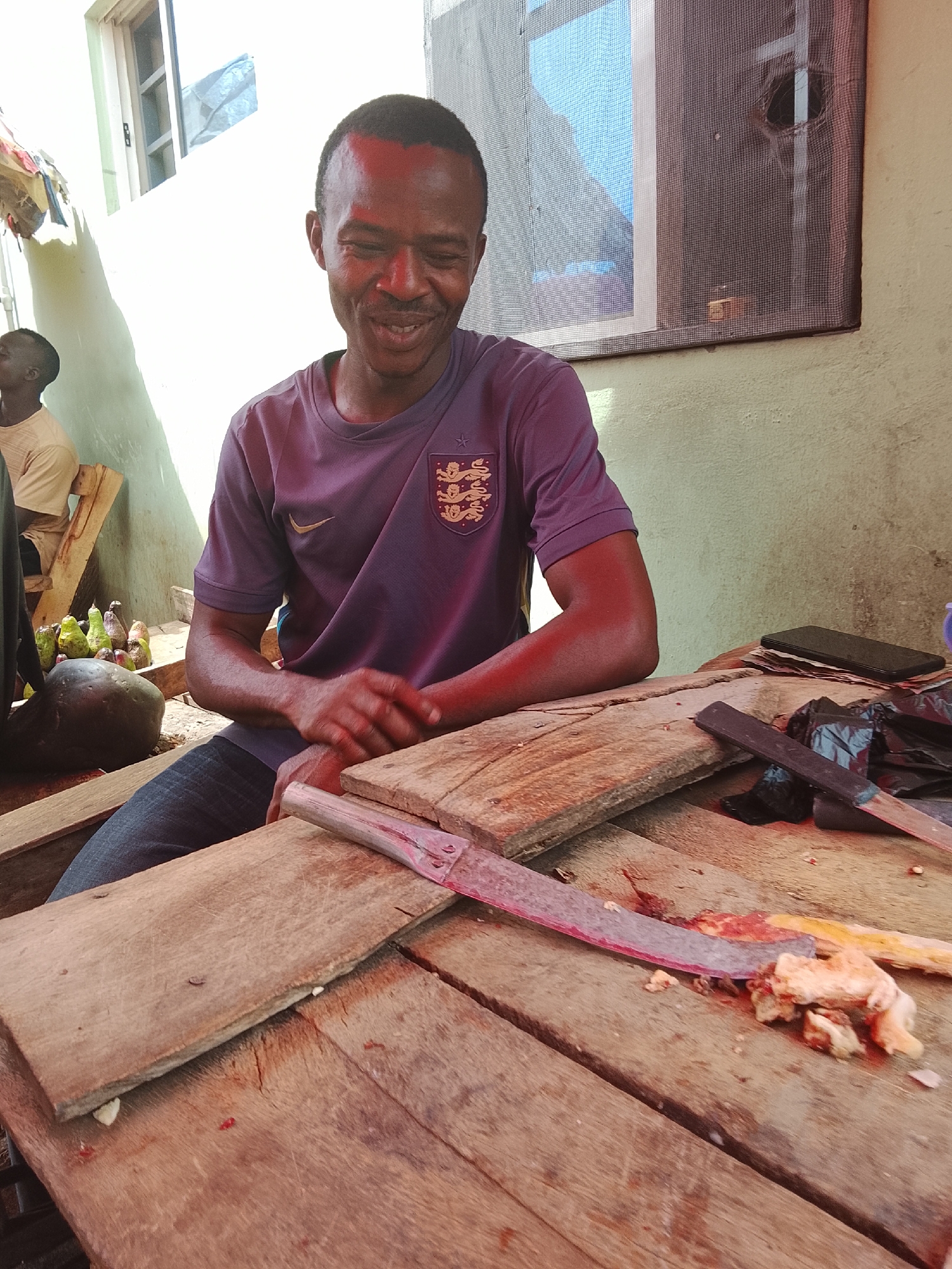 In Akwanga, Nigeria, a man sits at a wooden table. He is wearing a purple shirt with an emblem of three lions and a Nike logo, blue jeans, and has dark skin. The man is looking down and smiling. On the table in front of him is a large cleaver with what appears to be meat and bone remnants. A smaller dark object resembling a knife and a cell phone are also present on the table. To the man's left are a collection of green and brown items that appear to be gourds or other produce. Another individual is seated in the background. A window is above the man with screen and blinds. The overall environment is an outdoor workspace.