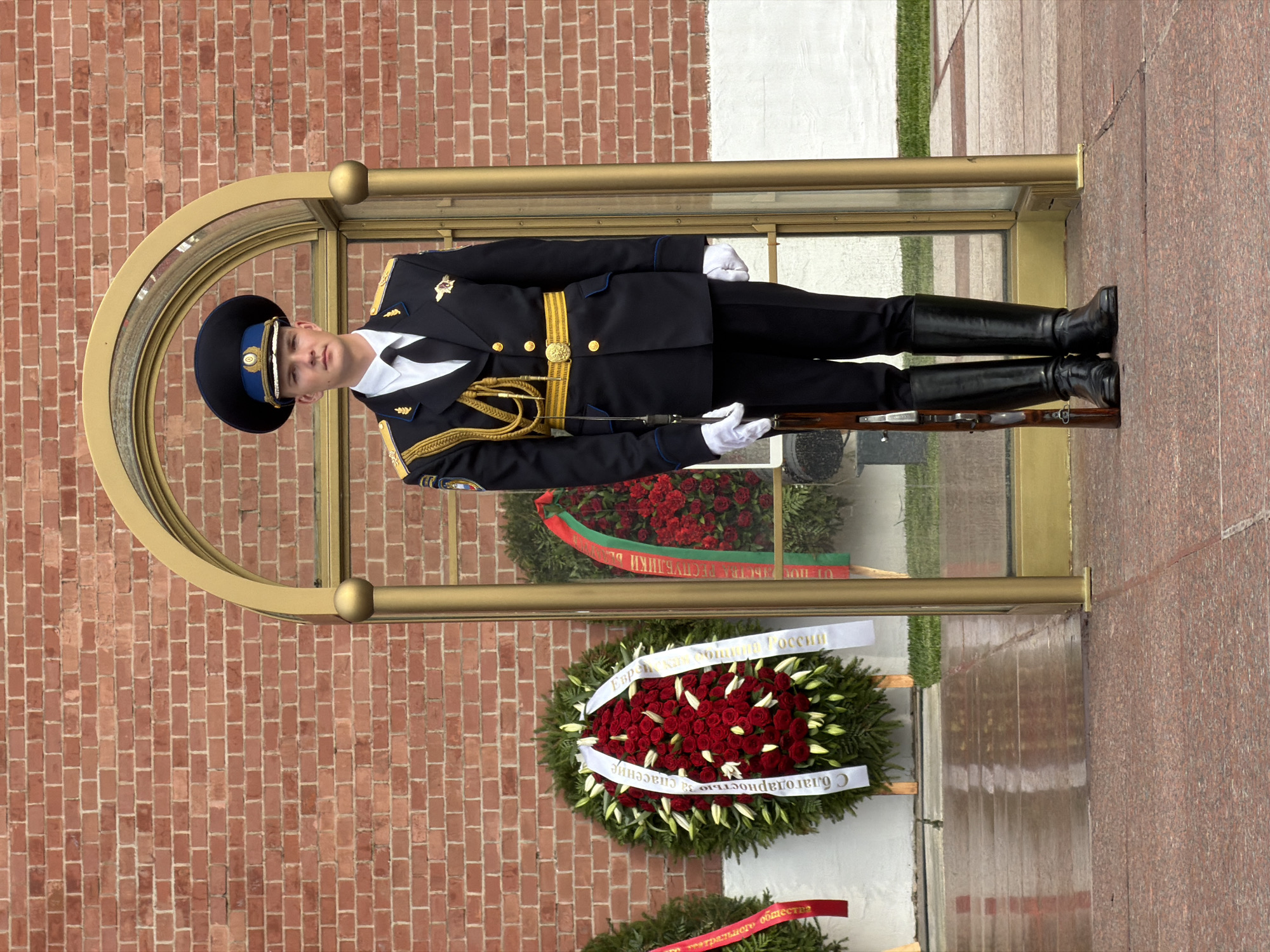 A member of the Presidential Regiment is standing guard at the Tomb of the Unknown Soldier in Alexander Garden, near the Kremlin wall in Moscow, Russia. The sentry is wearing a dark blue uniform with gold trim, a black cap, and black boots. He is holding a rifle. The Tomb is enclosed in a glass and gold frame. Wreaths are placed below the Tomb. The background shows a brick wall.