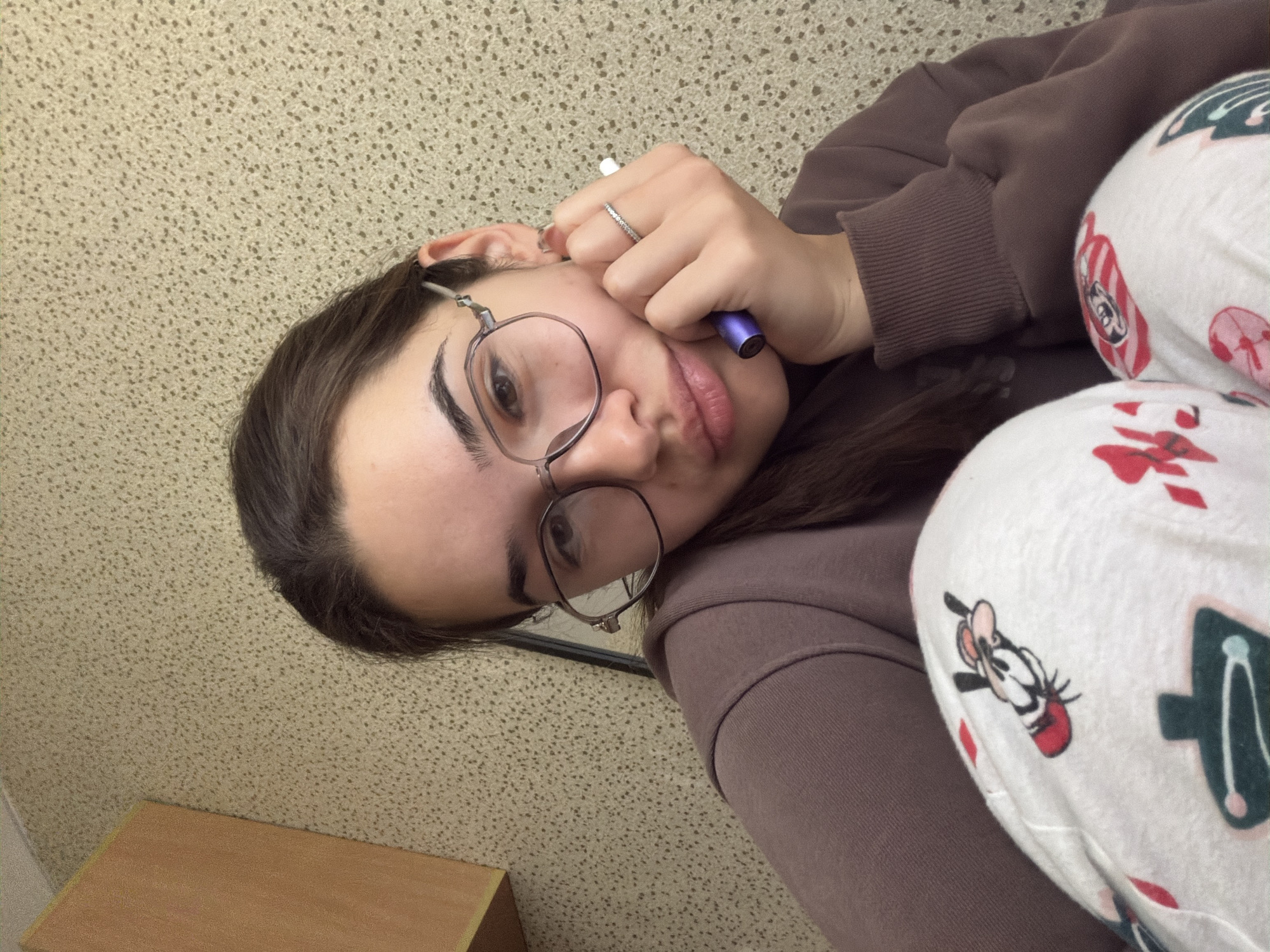 The image, oriented vertically, captures a close-up of a young woman lying on her side, looking directly towards the camera from a slightly elevated perspective. Her head is positioned towards the left side of the frame.
The woman has dark brown hair and wears clear-framed, slightly hexagonal glasses. Her eyebrows are dark and well-defined, and her lips are subtly pursed. She is dressed in a casual brown hooded sweatshirt or jumper and white pajama bottoms featuring colorful patterns, including a recognizable cartoon character that appears to be Pluto or Goofy from Disney, depicted with a red collar and hat.
Her right hand is raised near her face, holding a small, purple cylindrical object, likely a vape pen. A silver-toned ring adorned with small, clear stones is visible on her right ring finger.
The background consists primarily of a light beige or off-white wall with a fine, speckled or textured pattern, suggesting wallpaper or painted stucco. A small section of a light-colored wooden surface, possibly a desk or table, is visible in the lower left corner.
The woman's expression appears relaxed or thoughtful. The setting is indoors, with no specific visual cues indicating the time of day. No legible text is visible on any objects or clothing within the frame. The scene is set in Sofia, Bulgaria.