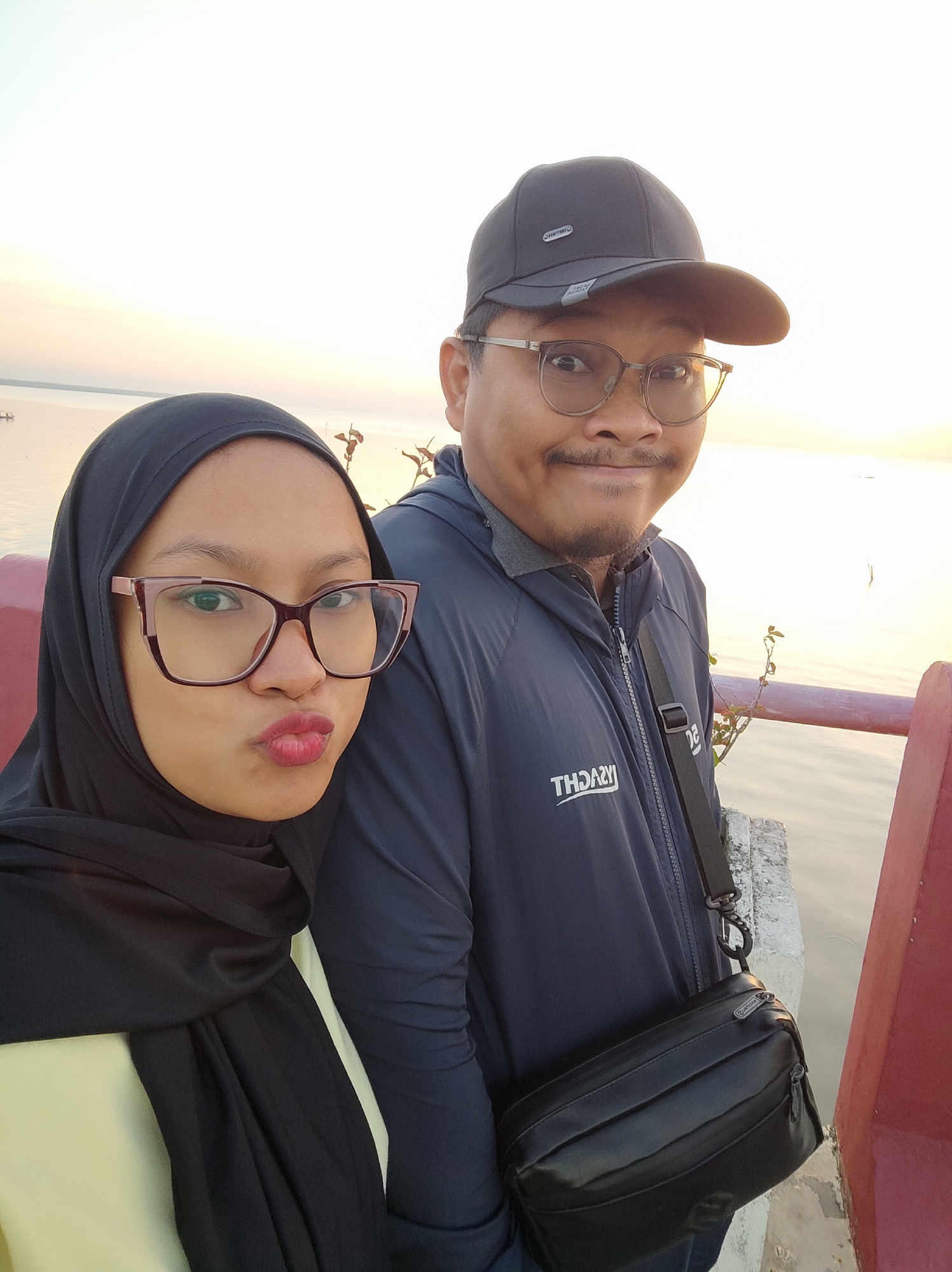The image is a selfie taken outdoors, likely at sunset given the warm light. Two individuals are visible. On the left, a woman wearing a black hijab and glasses pouts her lips. She is wearing a light-colored garment underneath the hijab. On the right, a man wearing a black baseball cap and glasses smiles. He is wearing a dark blue jacket with a zipper and a black crossbody bag. The background shows a body of water, possibly a river or lake, with a pale sky. A red railing is partially visible in the foreground on the right. The scene appears to be along a waterfront promenade or a similar public area. Based on the style of the people and the general ambiance, this could be a scenic spot in Surabaya, Indonesia, popular for evening strolls or capturing memories.