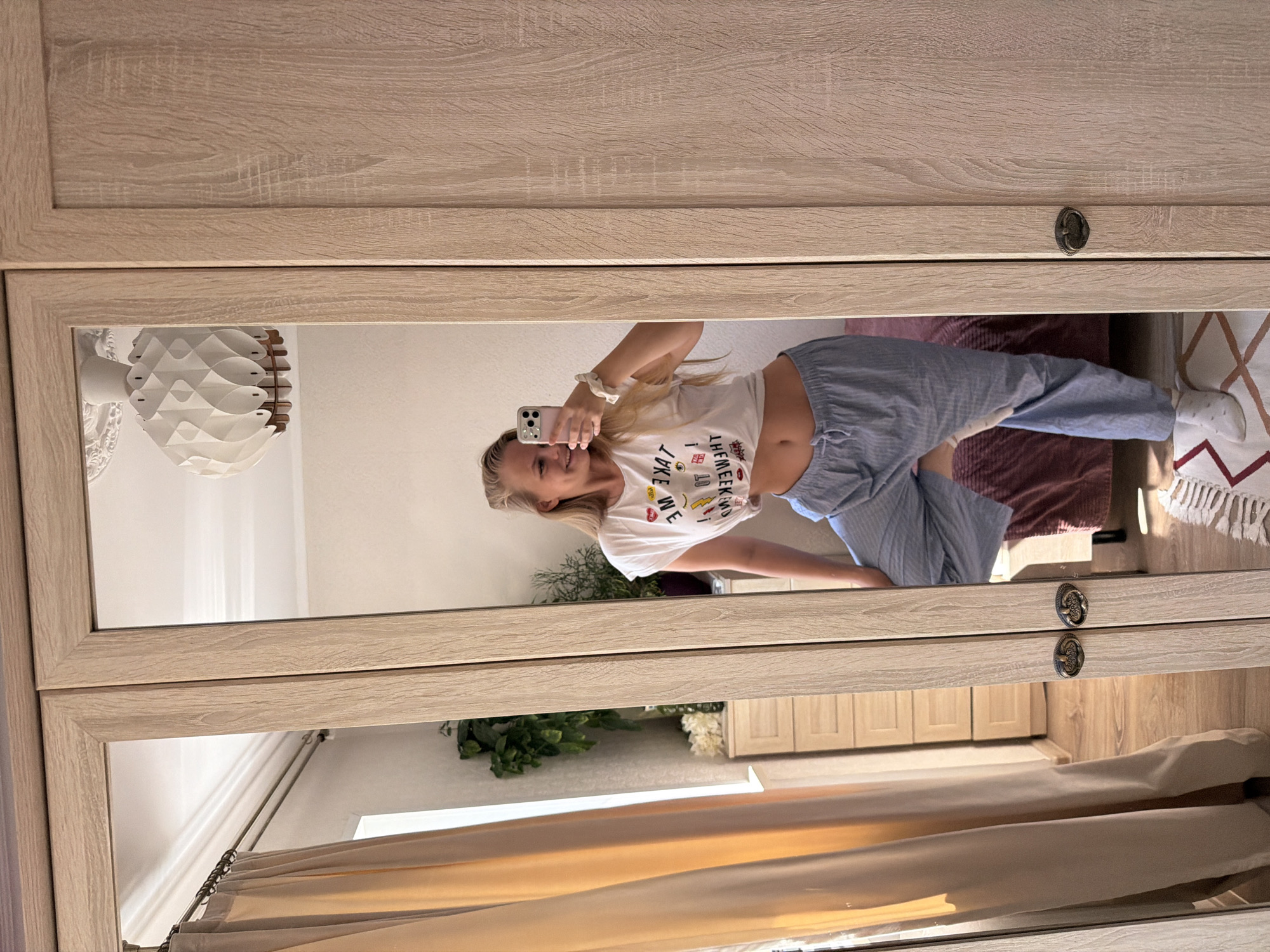 A young woman with blonde hair, wearing a white crop top and blue striped pajama pants, takes a mirror selfie in a bedroom. She is holding a pink smartphone in her right hand, and her left arm is bent with her hand on her hip. Behind her, a beige curtain is visible on the left, and a wooden dresser with several drawers is positioned against a wall. A large white geometric pendant lamp hangs from the ceiling, reflected in the mirror. The room appears to be located in an apartment in Chaikovsky, Russia, as indicated by the context of the prompt.