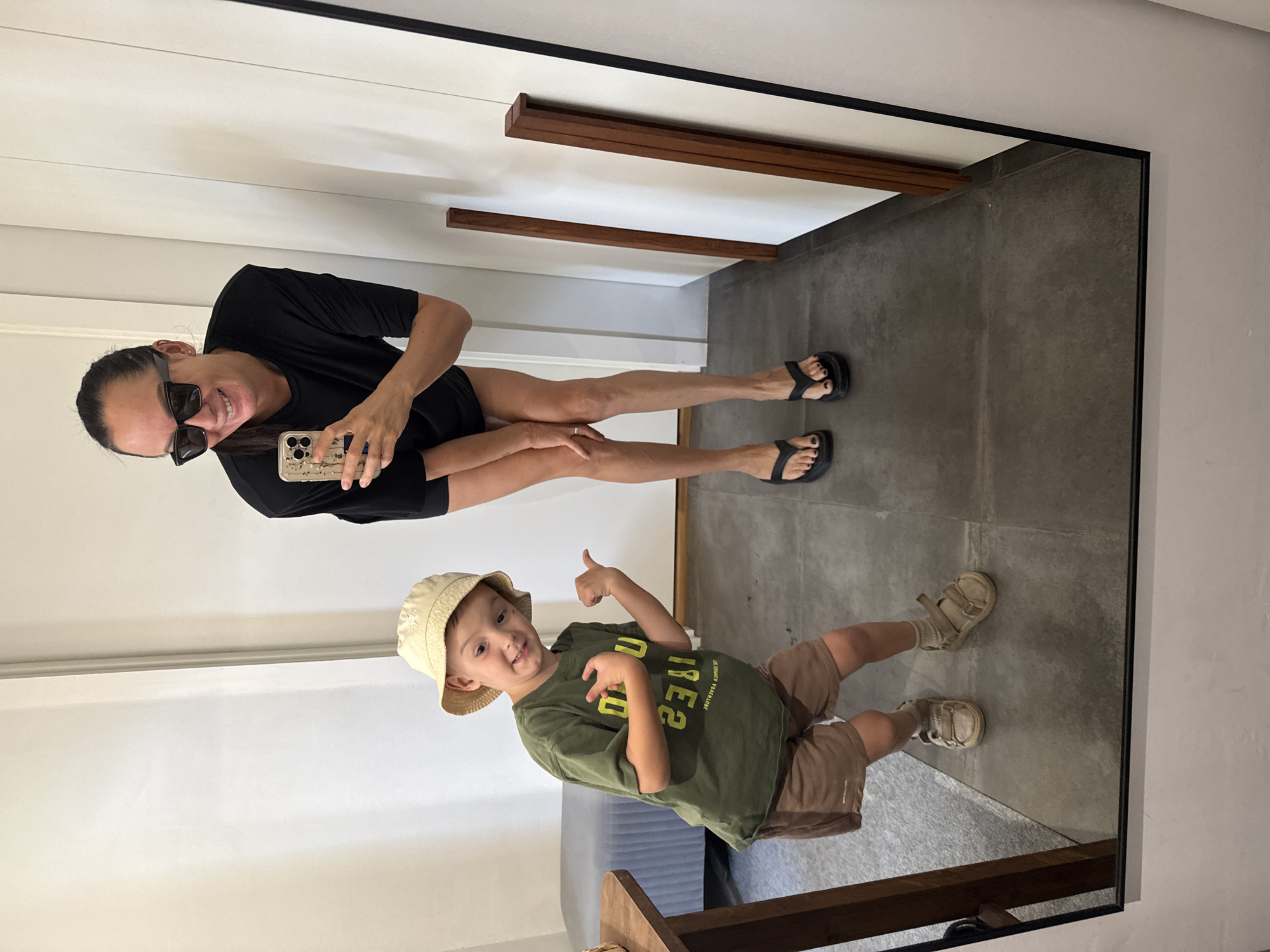 In a retail fitting room, a woman wearing sunglasses and a black top stands in front of a full-length mirror, holding a mobile phone to take a selfie. To her left, a young boy wearing a bucket hat, green t-shirt, and brown shorts poses playfully, gesturing towards himself and then to the side. The floor is tiled, and a wooden table is visible on the left. Wooden beams are attached to the wall behind the woman. There are no recognizable landmarks indicating the specific location within Chaikovsky, Russia.