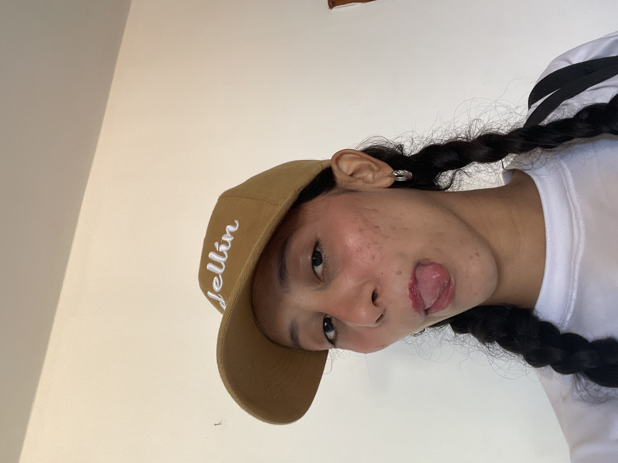 A young woman with dark hair in braids, wearing a tan baseball cap with white lettering that reads "Jellín", and a white t-shirt, is sticking her tongue out. She has a silver hoop earring in her visible ear. The background is a plain, light-colored wall, suggesting an indoor setting. The lighting appears to be natural, coming from the side, casting a slight shadow on the right side of her face. The image is taken from a close-up perspective, focusing on the woman's face and upper body. There are no elements in the image that provide a definitive location within the city of Porlamar, Venezuela.