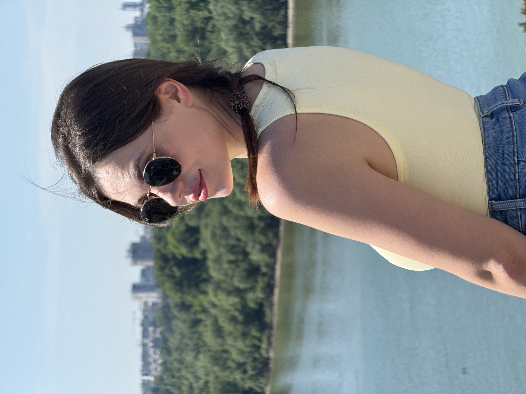 A woman with dark hair, wearing sunglasses, a light yellow top, and denim shorts is outdoors near a body of water and trees. The background shows a cityscape, suggesting the location is in an urban area with natural elements. It is daytime with clear skies.