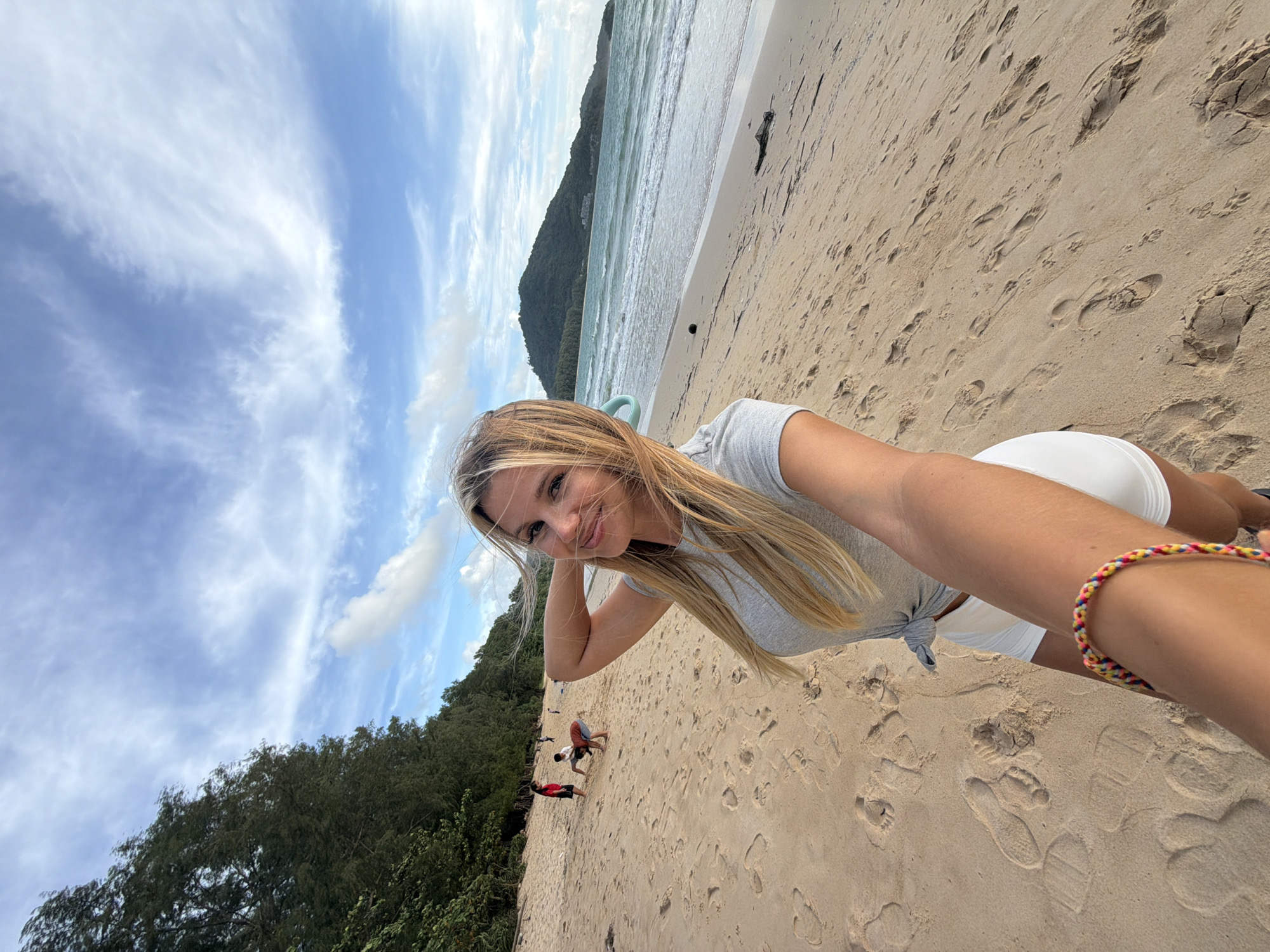 A blonde woman takes a selfie on a sandy beach in Sakhu, Thailand. She is wearing a grey t-shirt tied at the waist and white shorts. She is holding her phone in her right hand, which is extended towards the camera. Her left arm is bent at the elbow, with her hand resting on the back of her head. Her hair is blowing in the wind.
In the background, the sea meets the shore, with gentle waves rolling in. A lush green treeline runs along the edge of the beach, with a hill rising in the distance. A few people are visible in the mid-ground, walking and playing on the sand. The sky is blue with scattered white clouds. The sand is littered with footprints. A colorful bracelet is visible on the woman's left wrist.