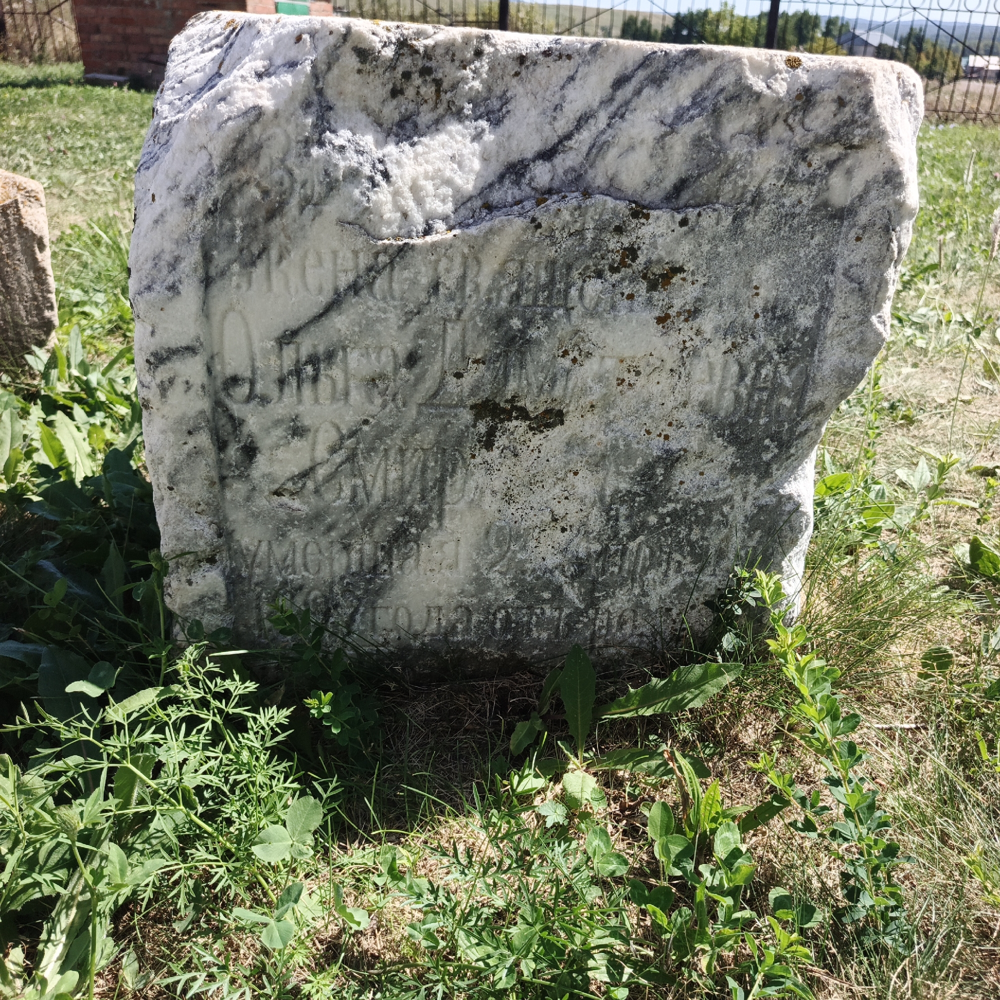 The image depicts a weathered, white marble tombstone with Cyrillic inscriptions, located outdoors in a grassy area. The tombstone is rough-hewn and shows signs of age, with dark veins running through the marble and patches of moss or lichen. The inscriptions are partially obscured by weathering and shadows but appear to be names and dates. The tombstone is surrounded by green grass and some weeds, with a blurred background that includes a fence, some vegetation, and structures that suggest a rural or cemetery setting. The lighting indicates it is daytime, with sunlight casting shadows. The location is identified as Тугустемир, Russia, suggesting a historical or memorial site within that region. There are no people visible in the image.