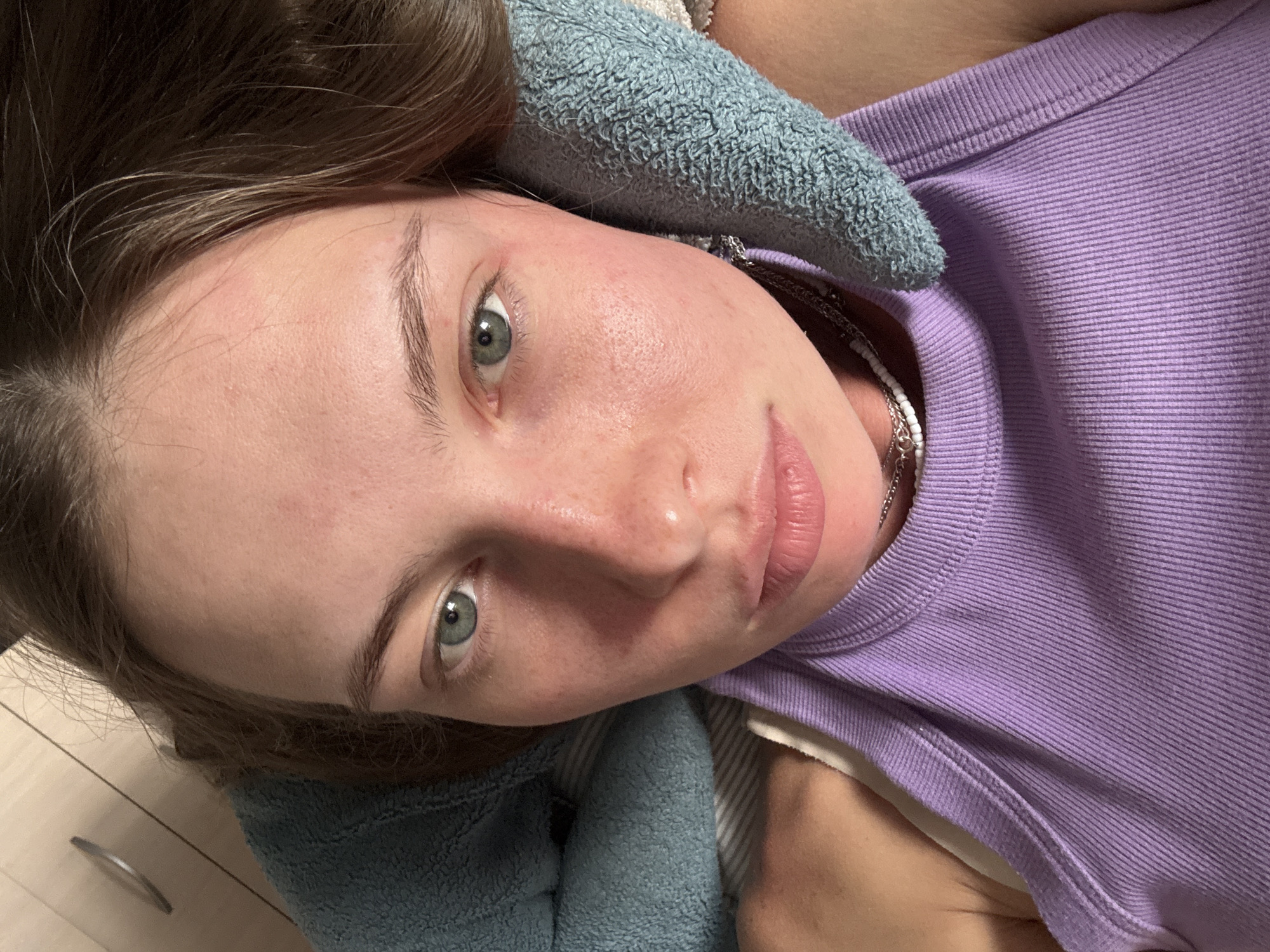 A close-up, high-angle shot captures a young woman lying down, looking directly into the camera. Her head is positioned to the left of the frame, and her body extends towards the right, suggesting a relaxed pose, possibly in bed. Her hair, a warm brown color, frames her face.
Her complexion is fair, with some minor redness visible on her cheeks and forehead. Her eyes are a striking blue-green, and her eyebrows are dark and well-defined. Her lips are naturally pink and slightly parted. She is wearing a light purple, ribbed tank top or camisole, visible on her right side, and a delicate silver chain necklace is peeking out from beneath the collar. A light blue, textured towel or blanket is draped around her head and neck, suggesting comfort or perhaps a recent hair wash.
The background is softly blurred, but hints of a wooden floor and a light-colored furniture piece with a silver handle are discernible in the lower left corner. The lighting appears soft and even, possibly indicating indoor lighting or an overcast day. Her expression is calm and neutral, with a slight hint of a smile. The image conveys a sense of intimacy and tranquility. There is no visible text in the image.