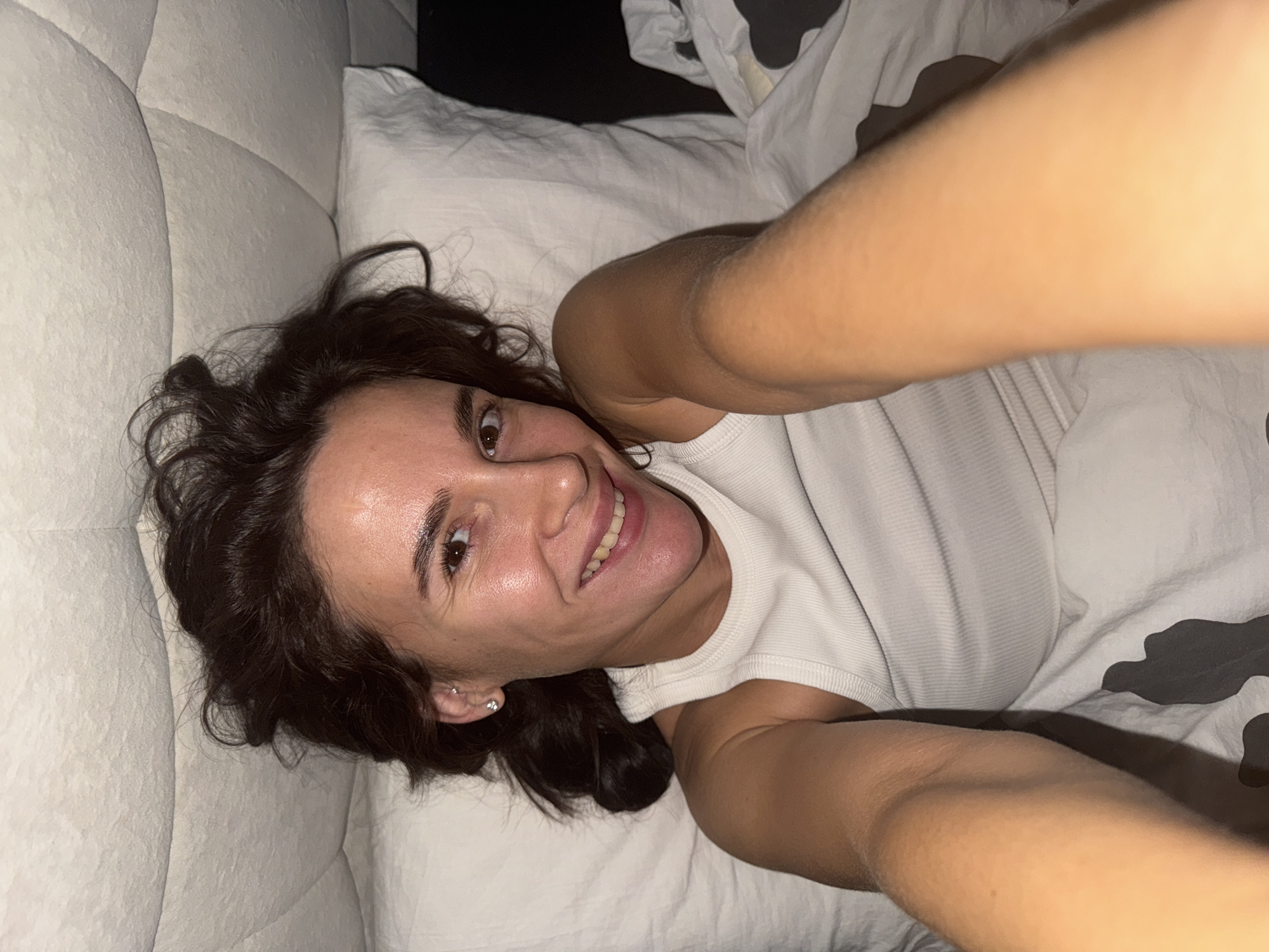 A young woman is lying in a bed. She is fair-skinned with dark brown hair, smiling at the camera and taking a selfie. She is wearing a white tank top and earrings. The bedding is white with a cow-patterned duvet or blanket on top of the white sheets. She is lying on a white tufted headboard. It is impossible to determine if this image was taken in the city of Chaikovsky, Russia from the image alone.