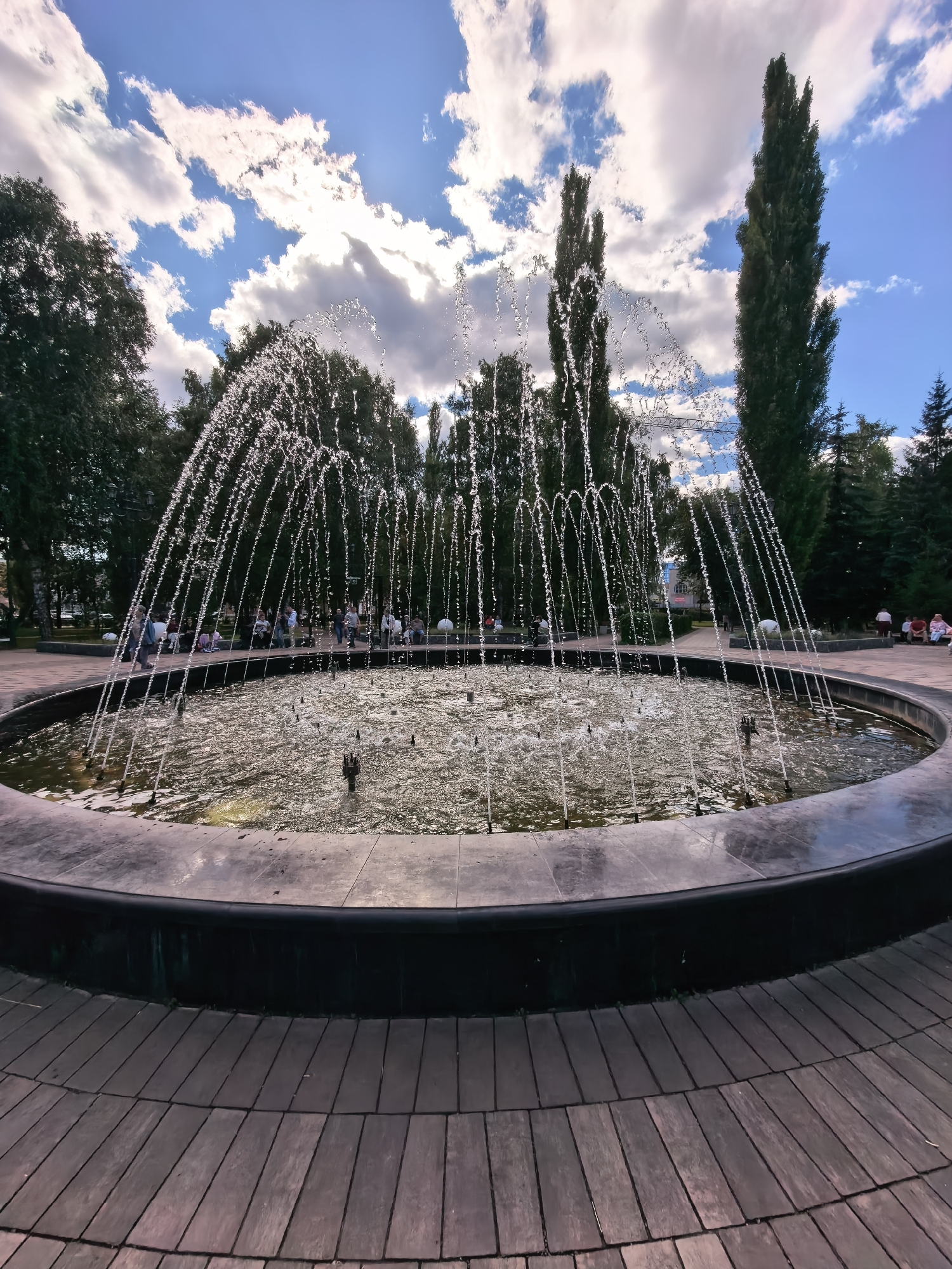The image shows a circular fountain in a park, likely located in the city of Sterlitamak, Russia. The fountain has multiple jets of water creating an arc shape. The water in the fountain is reflective and shows sunlight. The fountain is surrounded by a dark, circular border and a wooden deck made of planks. In the background, there are several people sitting on benches and walking around, suggesting it is a public space. Tall trees, including what appear to be cypress or poplar trees, are visible behind the fountain. The sky is blue with scattered white and gray clouds. The overall scene depicts a pleasant day in a park with a functioning fountain.