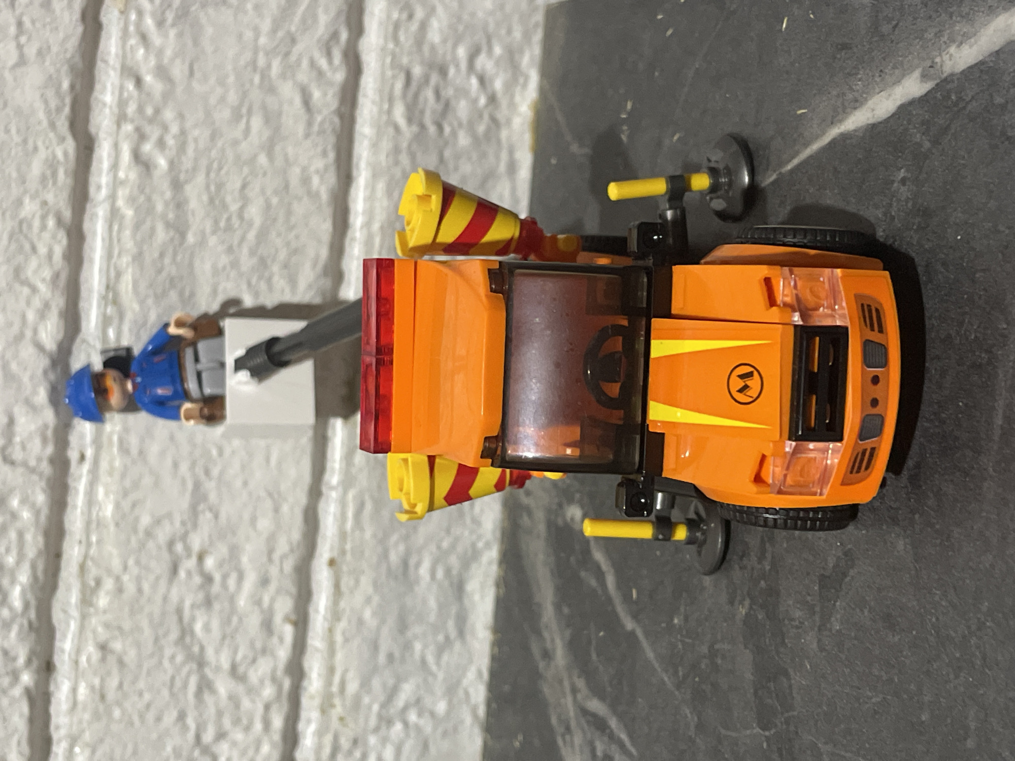 An orange toy construction vehicle is positioned on a dark grey, textured surface, possibly a table or floor. The vehicle has a red translucent light bar on top, a brown tinted windshield, and yellow accents on the front. Two yellow, cone-shaped objects with red and yellow stripes are mounted on either side of the rear of the vehicle. The vehicle has four black wheels. To the rear of the vehicle, a white platform supports a toy figure wearing a blue hard hat and a blue jacket over grey pants. The figure is holding a black pole that extends upwards and slightly to the right. The background consists of a white brick-patterned wall. The scene is likely indoors, and there is no indication of its location in Baranovka, Russia.