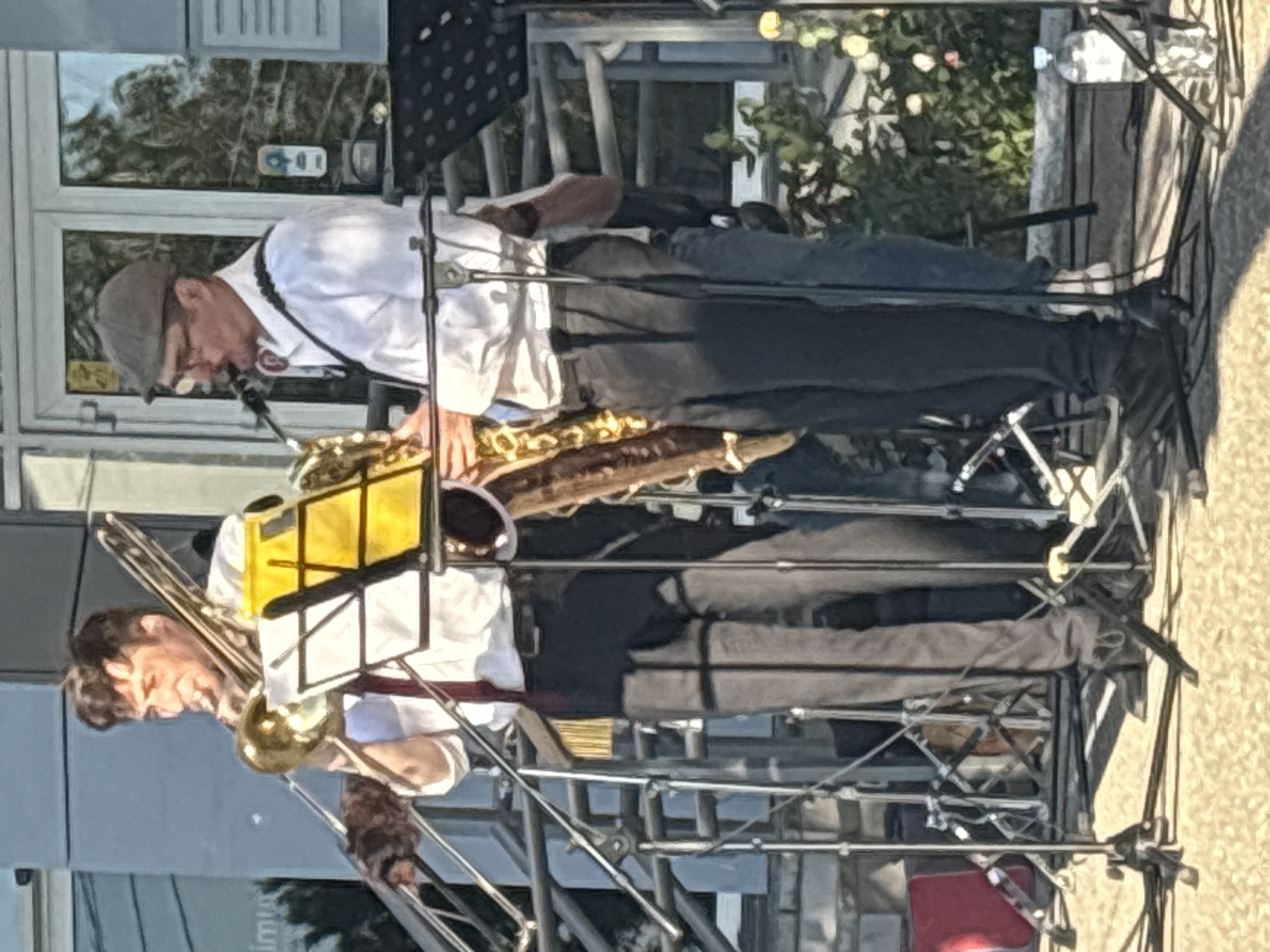 The image shows two men playing musical instruments outdoors. The man on the left is wearing a white shirt, black pants, and a grey flat cap. He is playing a saxophone. He appears to be singing into a microphone while playing. The man on the right is wearing a white shirt and black pants, and is playing a trombone. Both men are on a stage or platform with musical equipment. The scene appears to be a daytime event, possibly a performance or a street festival. The background shows a building with windows and some greenery. The overall mood seems to be lively, as suggested by the musicians' activity. The location context is Astrakhan, Russia. There is no visible text in the image.