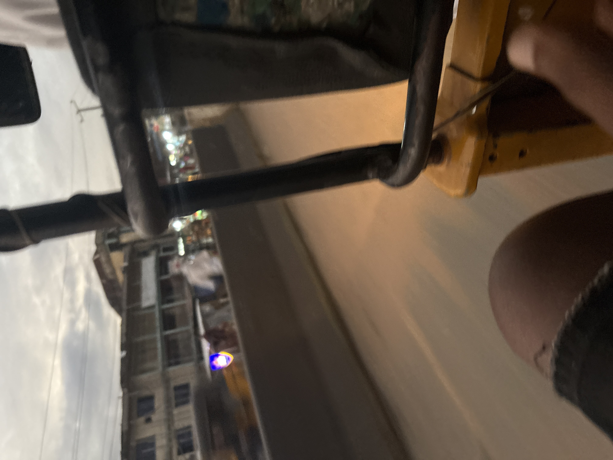 The image shows a view from a moving vehicle in Onitsha, Nigeria, likely a trishaw given the yellow bodywork and metal bar in the foreground. The scene captures an urban environment with buildings lining a street, illuminated by the warm glow of streetlights. The sky is overcast, suggesting it is either early morning or late afternoon.
In the background, several multi-story buildings with balconies and visible windows are present, typical of the architecture in Onitsha. Some of these buildings appear to be commercial establishments, with what seem to be shops or market stalls visible in the distance. The street itself is populated with blurred figures and vehicles, indicating the bustling nature of the city. One figure, possibly wearing protective gear, stands out amongst the crowd, though their identity and role are unclear due to the motion blur.
The foreground of the image is dominated by the interior of the vehicle, with a black metal railing and the yellow frame of the vehicle's body. A portion of a person's leg, wearing dark clothing, is visible on the left side, and a hand is seen on the right side, suggesting the perspective of a passenger or driver. A small, bright blue and yellow light is also visible, possibly a signal light or decoration. The overall impression is a snapshot of everyday life in a busy Nigerian city, captured from a dynamic vantage point.