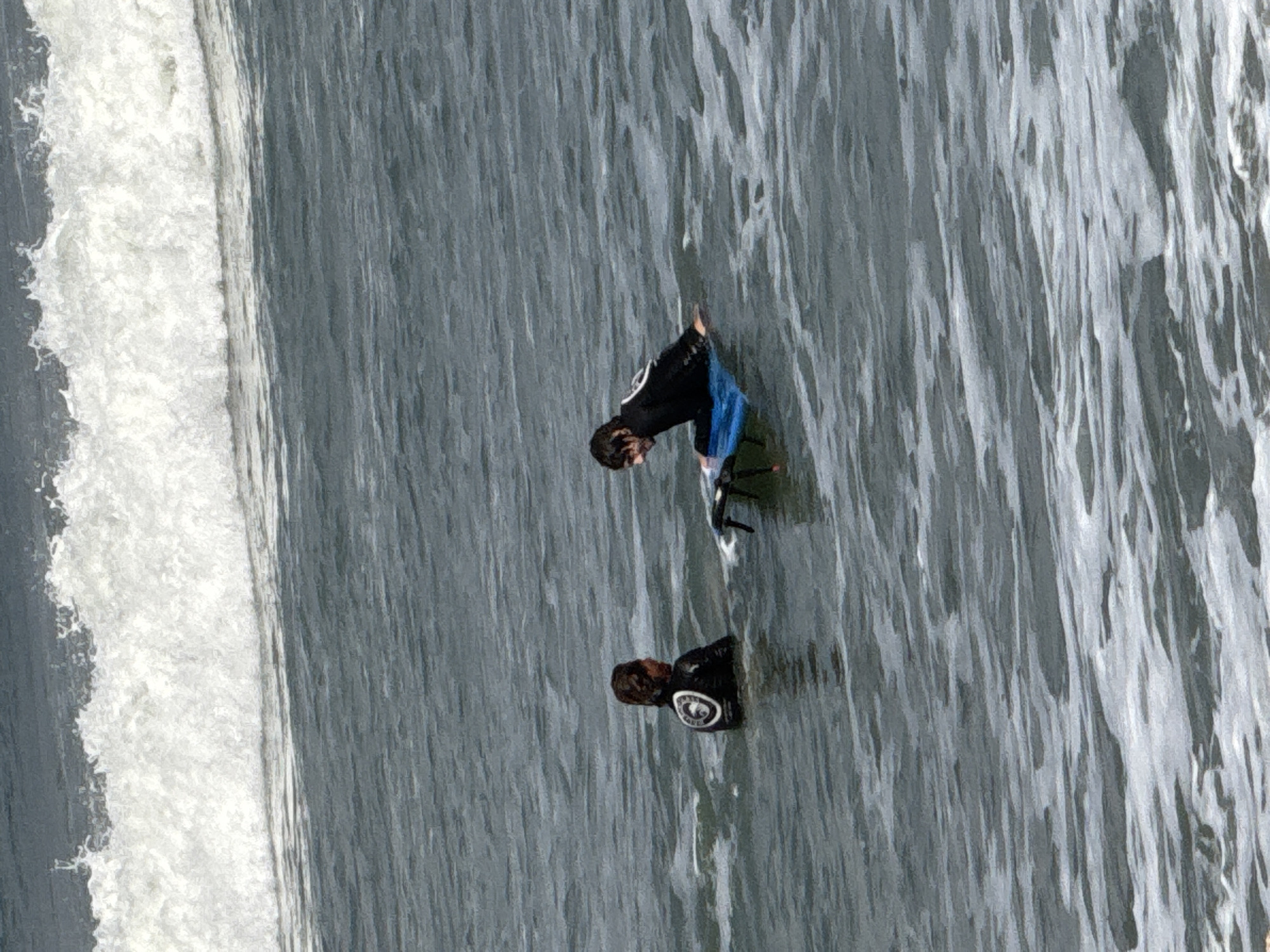 Two individuals in black wetsuits are in the ocean off the coast of Kuta, Indonesia. The person on the left is facing away from the camera, appearing to look towards the person on the right. The person on the right is sitting on a blue surfboard and is facing the person on the left. The ocean is choppy with white waves in the background.