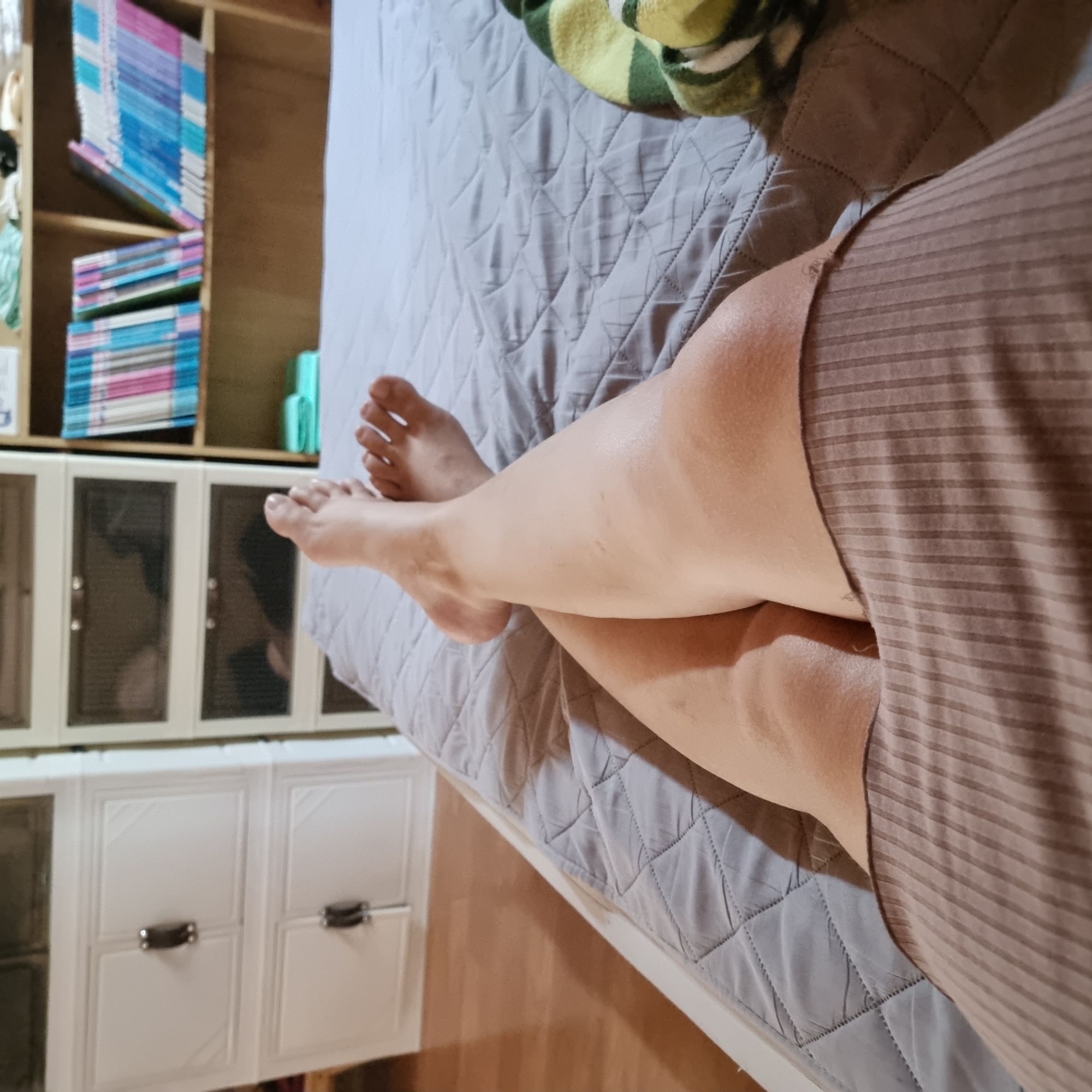 A person's legs are crossed and resting on a gray quilted bedspread in a room. The person is wearing a brown ribbed garment around their waist. To the left of the bed is a white cabinet with two drawers. To the right of the bed is a wooden bookshelf filled with books and folded green and white towels. The floor is made of light-colored wood. The room appears to be in Dunnae-myeon, South Korea.