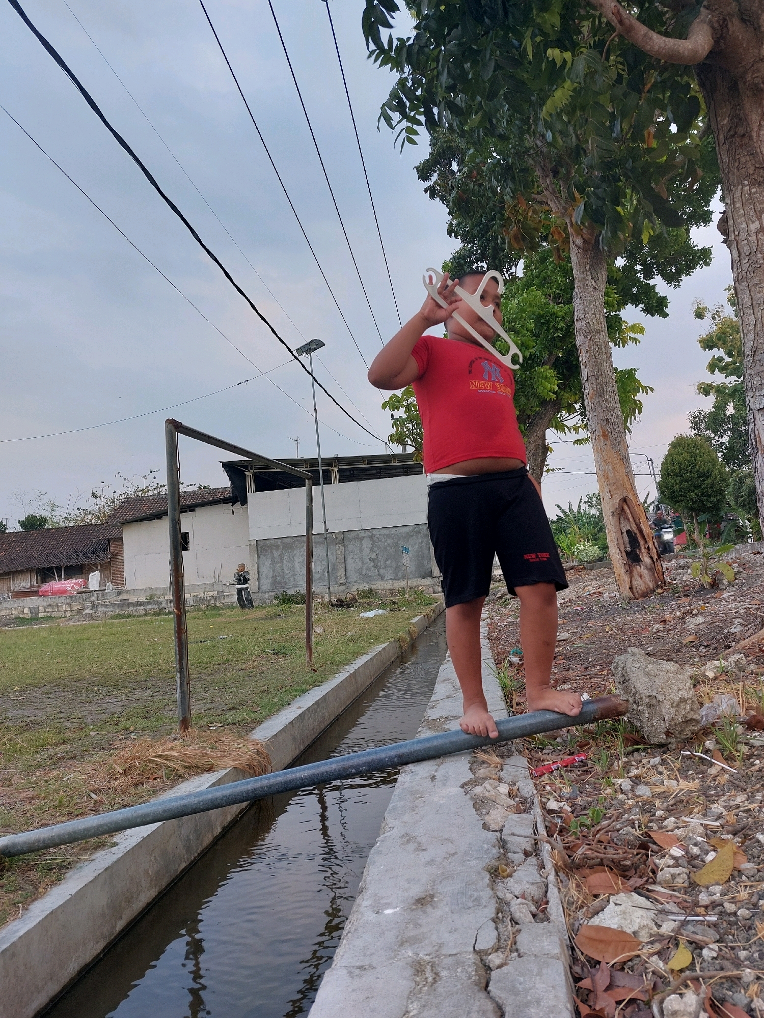 In Sarangan, Indonesia, a boy wearing a red t-shirt and black shorts stands on a metal pipe that spans a narrow concrete channel filled with water. The boy is holding two white hangers up to his face. The environment is outdoors, with trees and vegetation visible. In the background, there are buildings, a fence, and power lines. The sky is overcast. The scene suggests a casual, possibly playful moment.