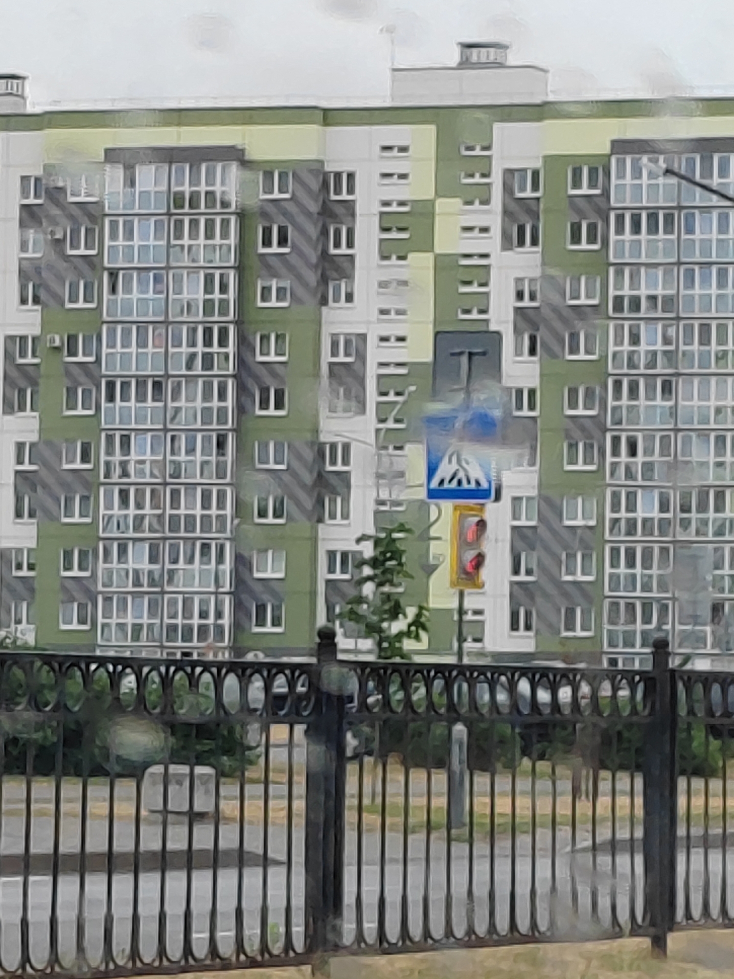 The image shows a modern apartment building with a checkerboard facade of light green and grey panels, accented with white window frames. The building has multiple floors and rows of windows, suggesting it is a residential complex. In the foreground, a black ornamental metal fence runs across the bottom of the frame, obscuring a view of a sidewalk and street. Behind the fence, there are trees and greenery. A pedestrian crossing sign and a traffic light with a red signal are visible on the right side of the apartment building, indicating a street intersection. The sky above is overcast and appears grey. The overall scene depicts an urban residential area.
