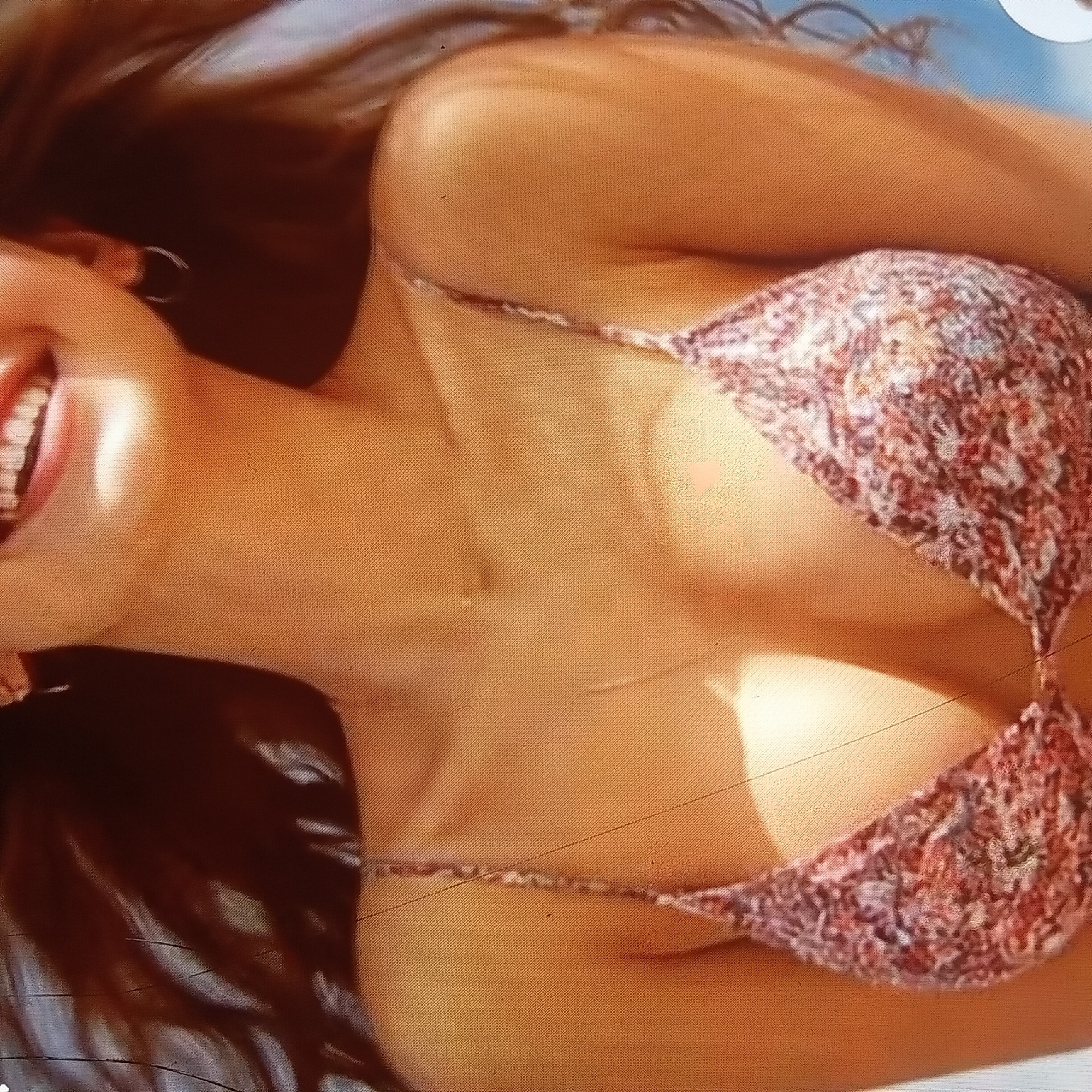 The image shows a close-up of a woman's torso. She is wearing a patterned bikini top. Her skin appears tanned, and she has long, dark hair that is out of focus. The background is blurred, but appears to be outdoors, possibly near water, given the light blue color. There are no discernible landmarks or features that would indicate a specific location in Almaty, Kazakhstan. The scene is brightly lit, suggesting it may be daytime.