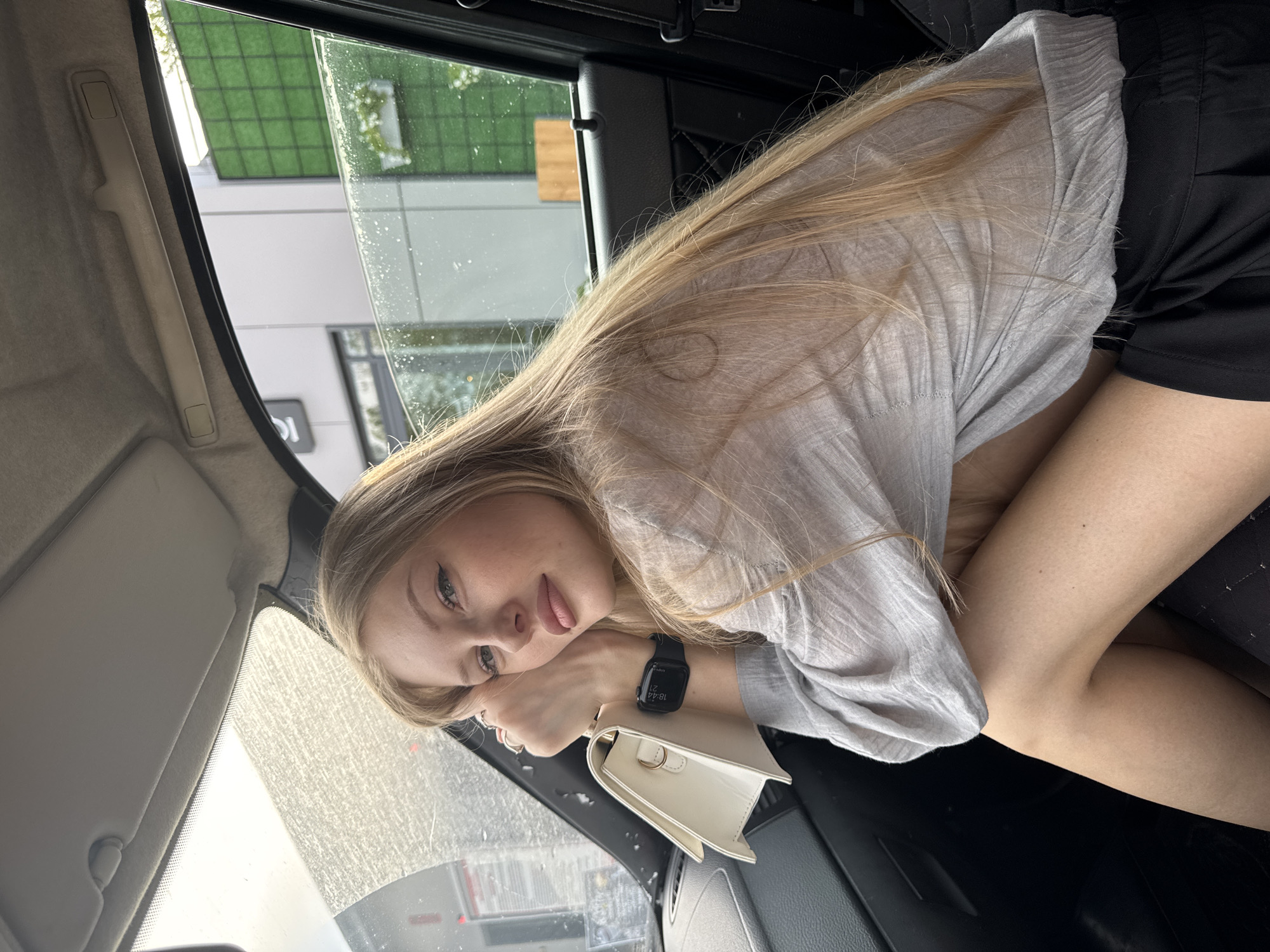 A young woman with long blonde hair sits in the passenger seat of a car, looking towards the camera. She is wearing a light gray, sheer long-sleeved shirt and black shorts. She holds a white handbag in her left arm, which rests on her knee. A black smartwatch is visible on her left wrist. The car interior is visible, including the dashboard and the car door. Through the car window, a building with green vertical panels and some foliage is visible. The weather appears to be overcast, with water droplets on the car window. The scene is set in a city.