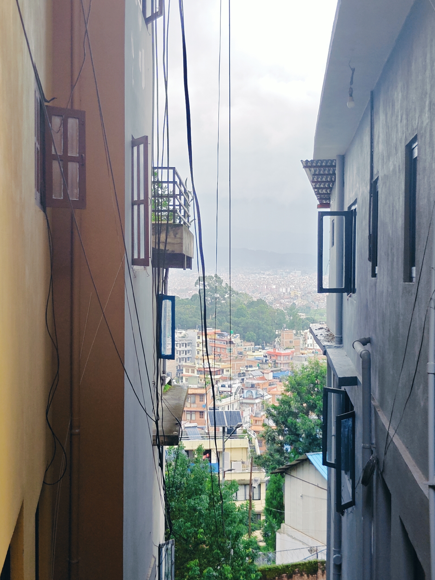 The image is a vertical shot of a narrow alleyway between two buildings, overlooking a densely populated cityscape, likely in Kirtipur, Nepal. The buildings on either side are close together, with numerous windows and balconies visible. Electrical wires are strung haphazardly across the scene, creating a tangled web overhead. The building on the left is painted yellow and brown, with a dark brown window frame and white-paned glass. The building on the right is a light gray concrete structure with multiple windows, some open and some closed, revealing dark frames. One of these windows has a blue frame. In the background, a sprawling city with a mix of architectural styles, including red-tiled roofs and multi-story buildings, stretches out towards a hazy, mountainous horizon. Lush green trees are interspersed throughout the urban landscape. The sky is overcast, suggesting a cloudy or possibly rainy day. The scene captures the characteristic urban density and infrastructure of a developing city.