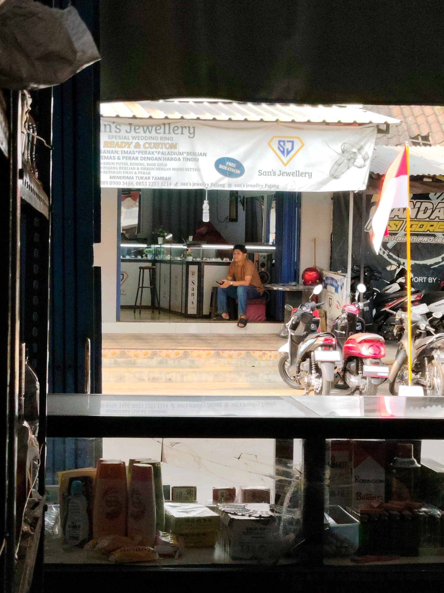 In Dampit, Indonesia, a jewelry store called "San's Jewellery" is captured. A white banner with "San's Jewellery" written on it hangs above the entrance, featuring a logo with a stylized diamond. A man in a brown shirt and blue jeans sits on a red cushion outside the store, looking at his phone. Motorcycles are parked in front of the store. Inside, displays of jewelry are visible behind glass counters, with a wooden stool in the foreground. The store is open, with its entrance revealing the interior. The scene is set on a street, with the sun illuminating the area.