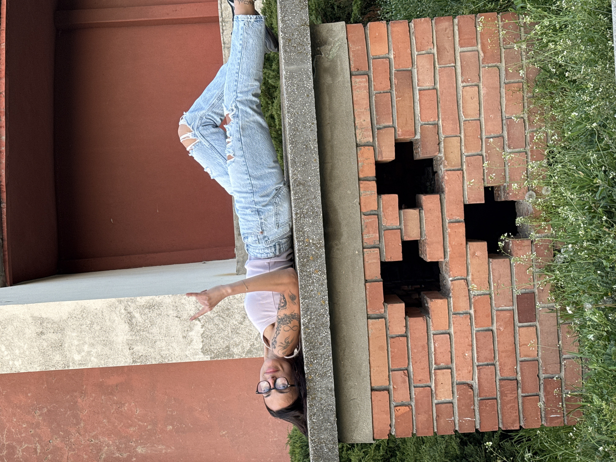 A woman in ripped jeans, a light-colored tank top, and glasses stands upside down, likely on a ledge. She has tattoos on her upper arm. She is positioned near a brick structure with a noticeable cutout. The scene appears to be outdoors, with greenery visible nearby. There is no information in the image to determine if the photo was taken in Pleven, Bulgaria.