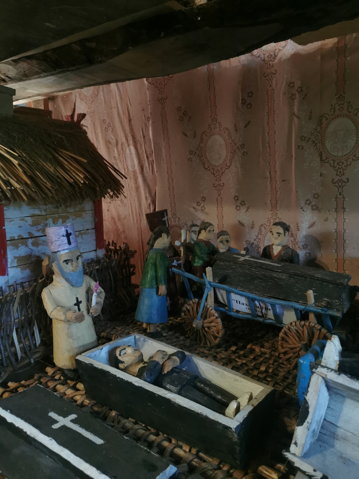 The image shows a diorama depicting a funeral scene. In the foreground, two wooden coffins are visible. The left coffin is black with a white cross painted on its lid, and the right coffin is open, revealing a wooden effigy of a deceased person lying inside. A wooden figure dressed as a priest, wearing a light-colored robe and a pink hat with a cross, stands to the left of the open coffin, holding a pink candle. Behind the coffins, a group of wooden figures, seemingly mourners, stand around a larger, dark-colored coffin mounted on a blue cart with woven wheels. The cart has a sign with text, part of which reads "Пахаванне" (Funeral). To the left of the scene is a small wooden structure with a thatched roof and weathered walls. The background features a pink fabric with an ornate, damask-like pattern. The overall setting suggests a representation of a traditional funeral in a rural Belarusian village, potentially in the Шчарчоўскі сельскі Савет region of Belarus.