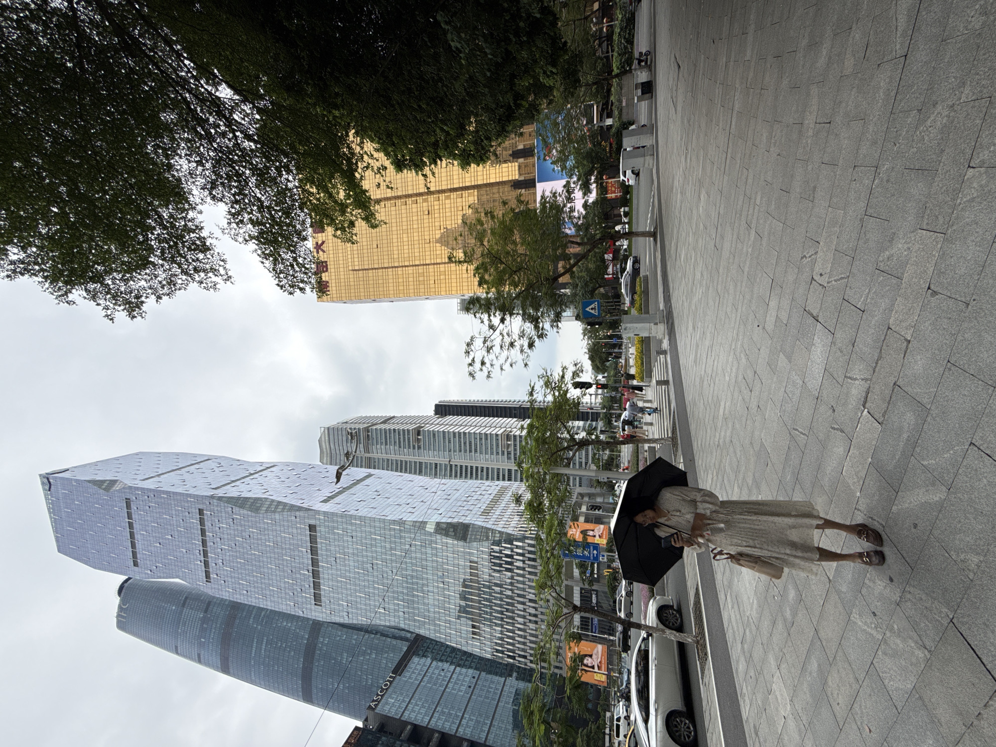 In the Tianhe District of China, a woman stands on a paved sidewalk, holding an open black umbrella and a smartphone. She wears a long-sleeved, floral-patterned dress and brown loafers. A beige tote bag hangs from her left shoulder. Behind her, a street is lined with modern skyscrapers, including a distinct golden building. Cars and pedestrians are visible on the street and across the intersection. Trees and greenery are interspersed among the urban structures. The sky is overcast.