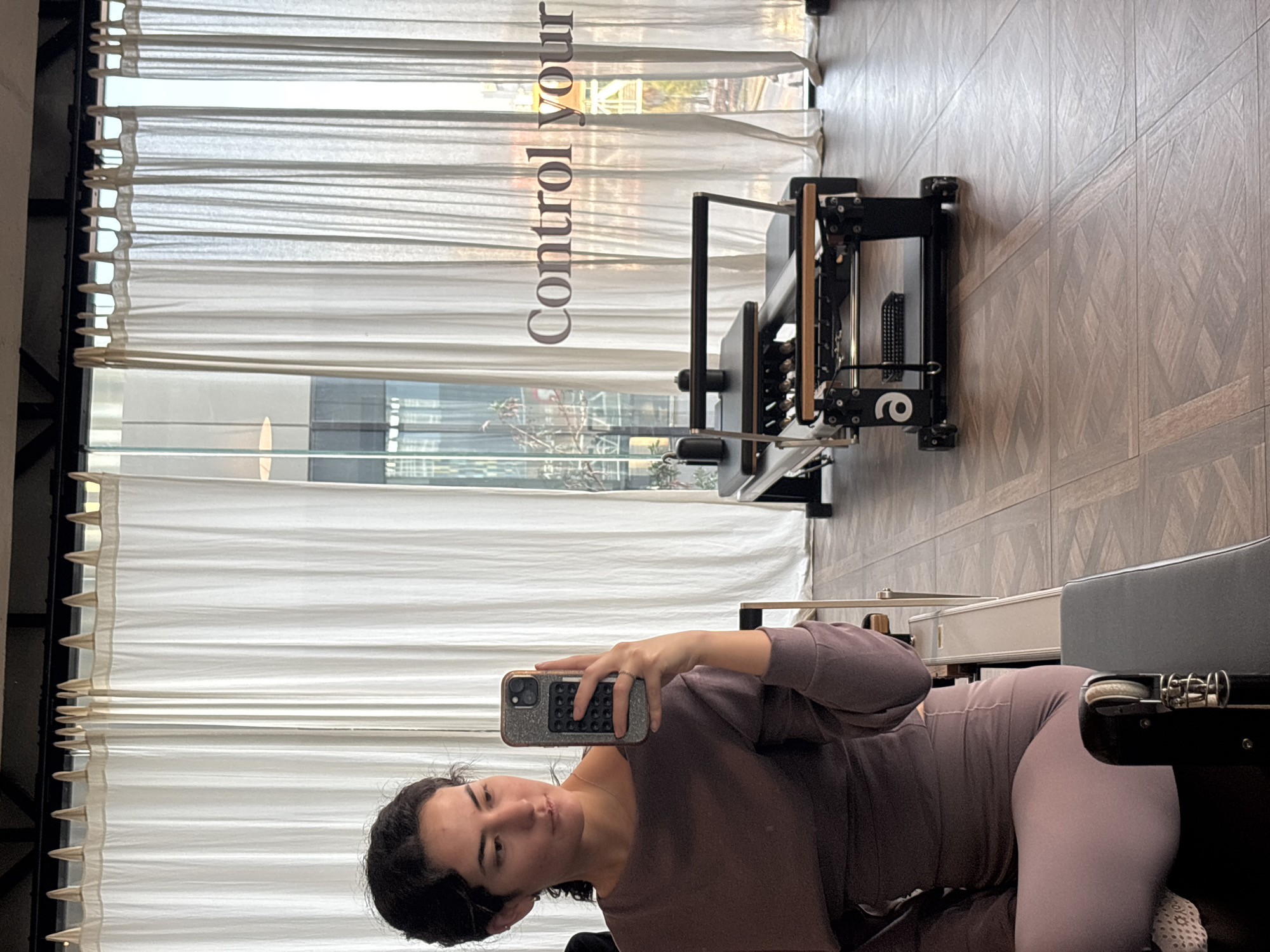 A woman is taking a selfie in what appears to be a pilates studio. She is lying on a pilates machine. The studio has light-colored wooden floors and large windows covered with white blinds. The words "Control Your" are visible on the window or blinds. It is not possible to confirm the location within the city of San Bernardino Tlaxcalancingo, Mexico from this image.